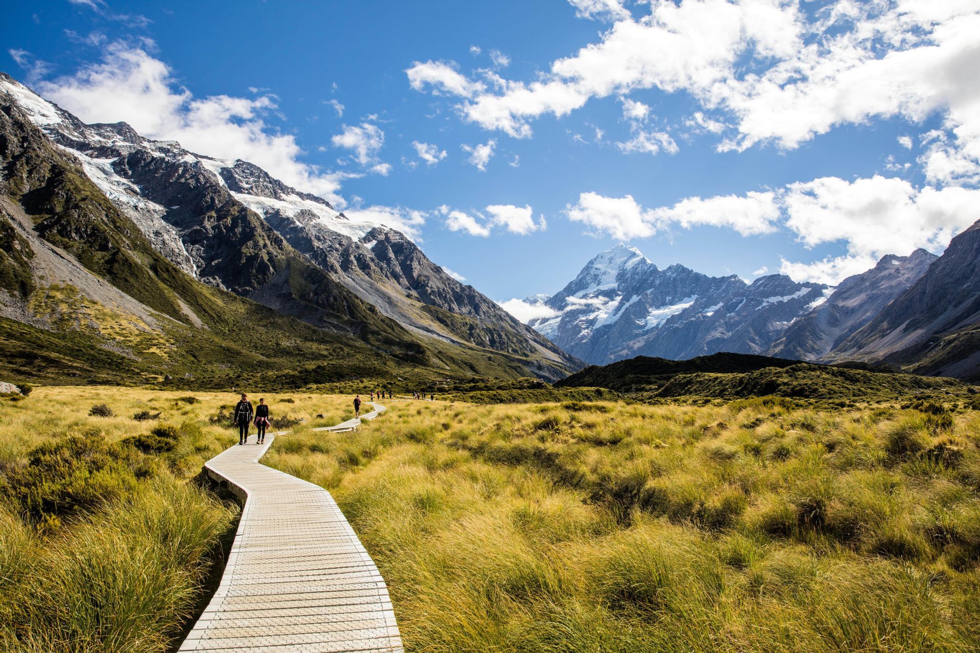 Un voyage de groupe WeRoad se promène sur une passerelle en bois, traversant une vallée verdoyante, avec des montagnes enneigées en fond.