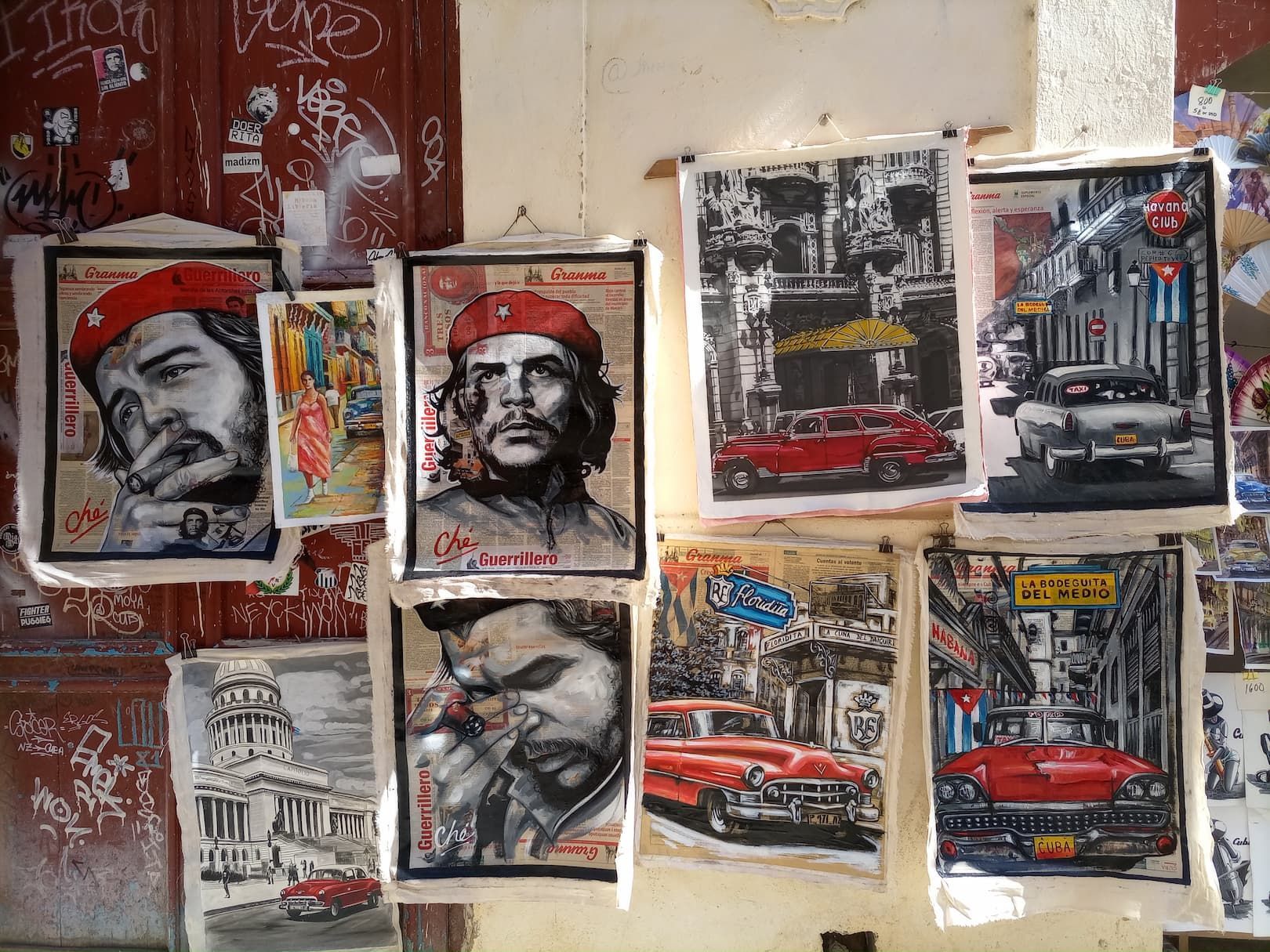 Une collection de tableaux thématiques cubains, mettant en scène Che Guevara et des voitures classiques, orne un mur couvert de graffitis.