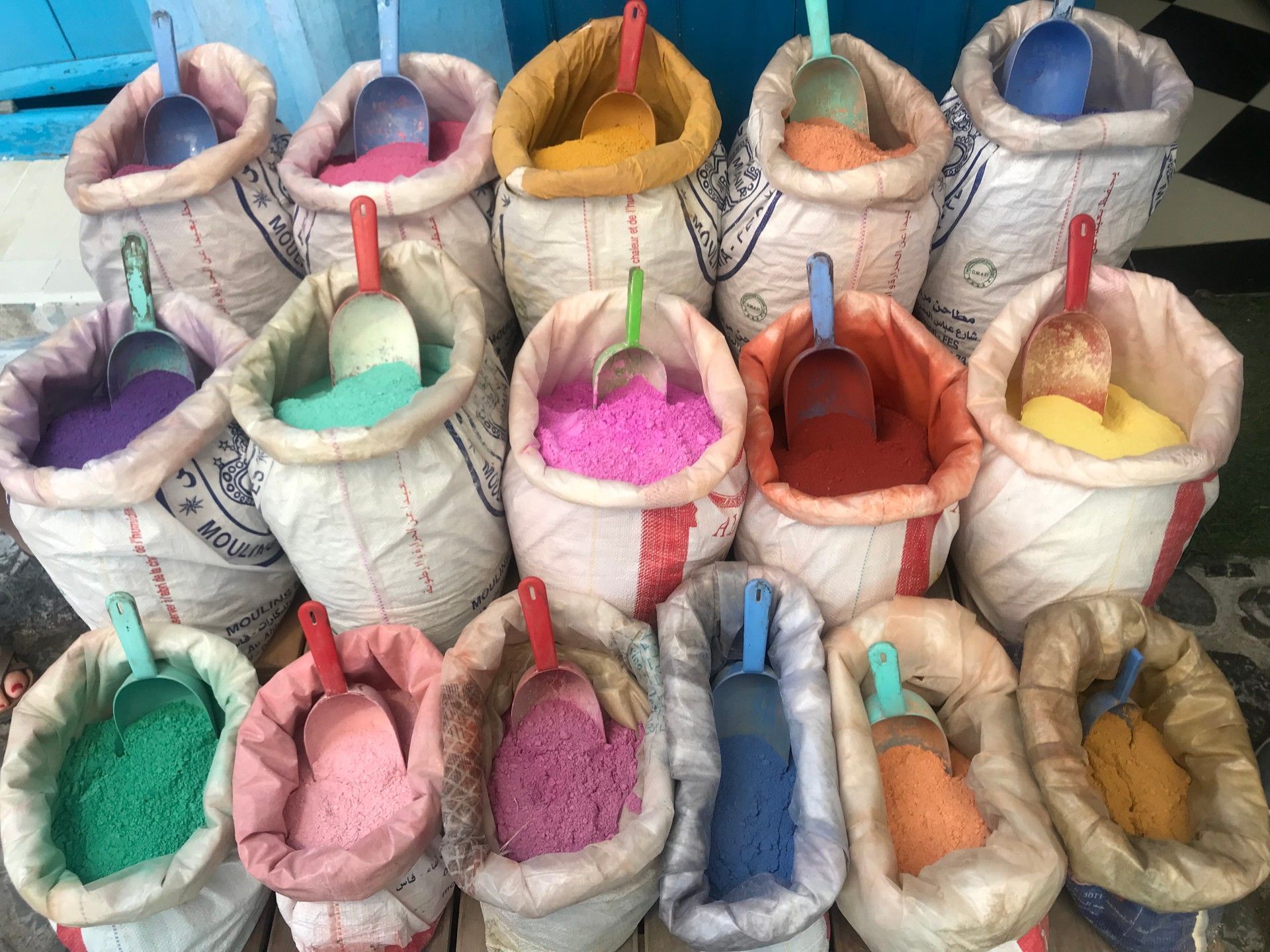 A collection of open white sacks filled with brightly colored powdered pigments, each with a plastic scoop inside.