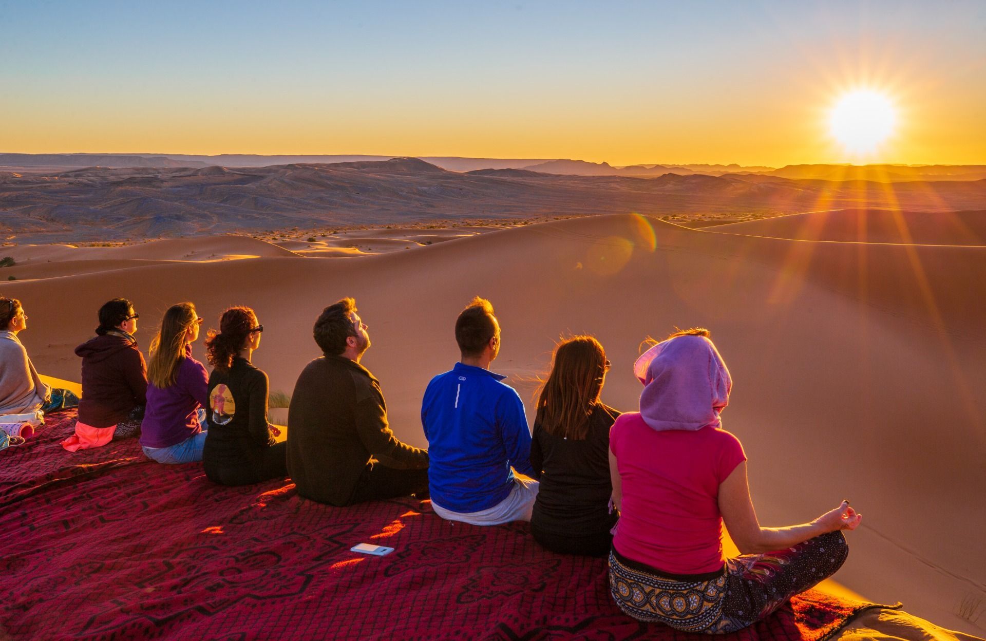 A WeRoad group trip sits on a blanket on a sand dune, watching the sunset over a vast desert landscape.
