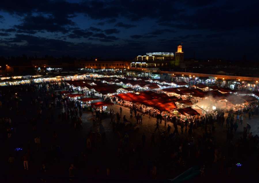 A bustling outdoor market square at night, filled with crowds of people among rows of illuminated stalls with red awnings.