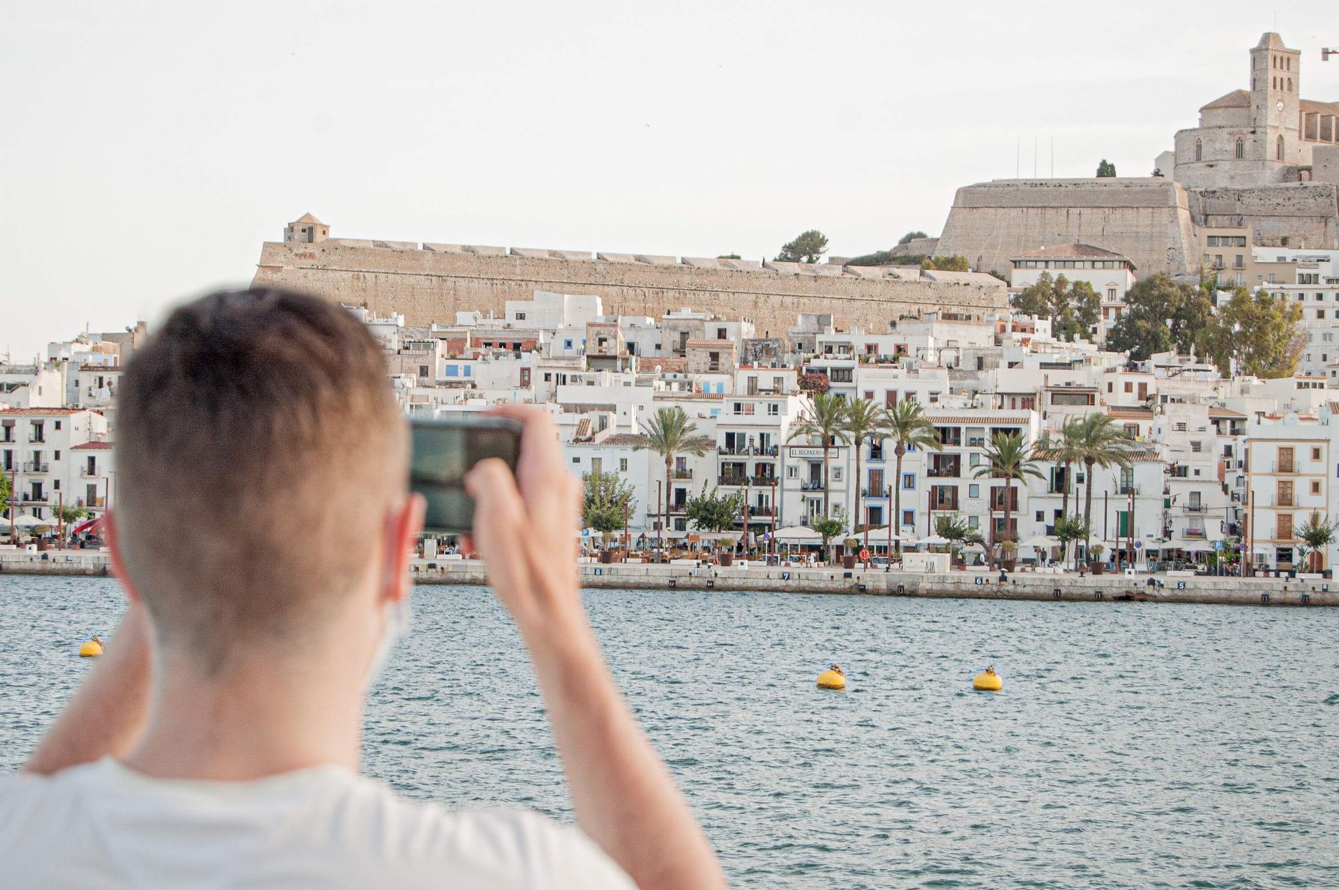 A person, seen from behind, takes a photo with a smartphone of a coastal town with white buildings and a hilltop fortress.