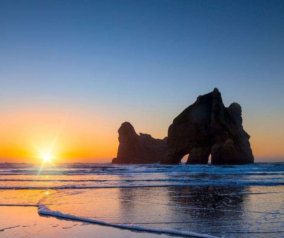The sun sets over the ocean, silhouetting a large rock formation with arches as gentle waves wash onto a sandy beach.
