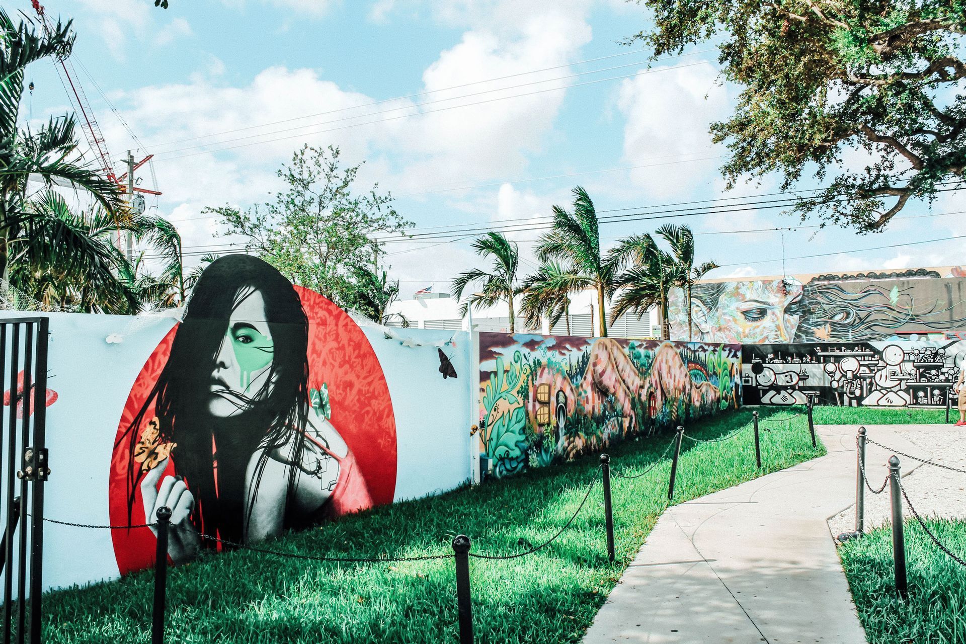 Un muro exterior cubierto de arte callejero colorido, con un gran mural de una mujer, junto a un césped verde y un camino bajo un cielo azul.