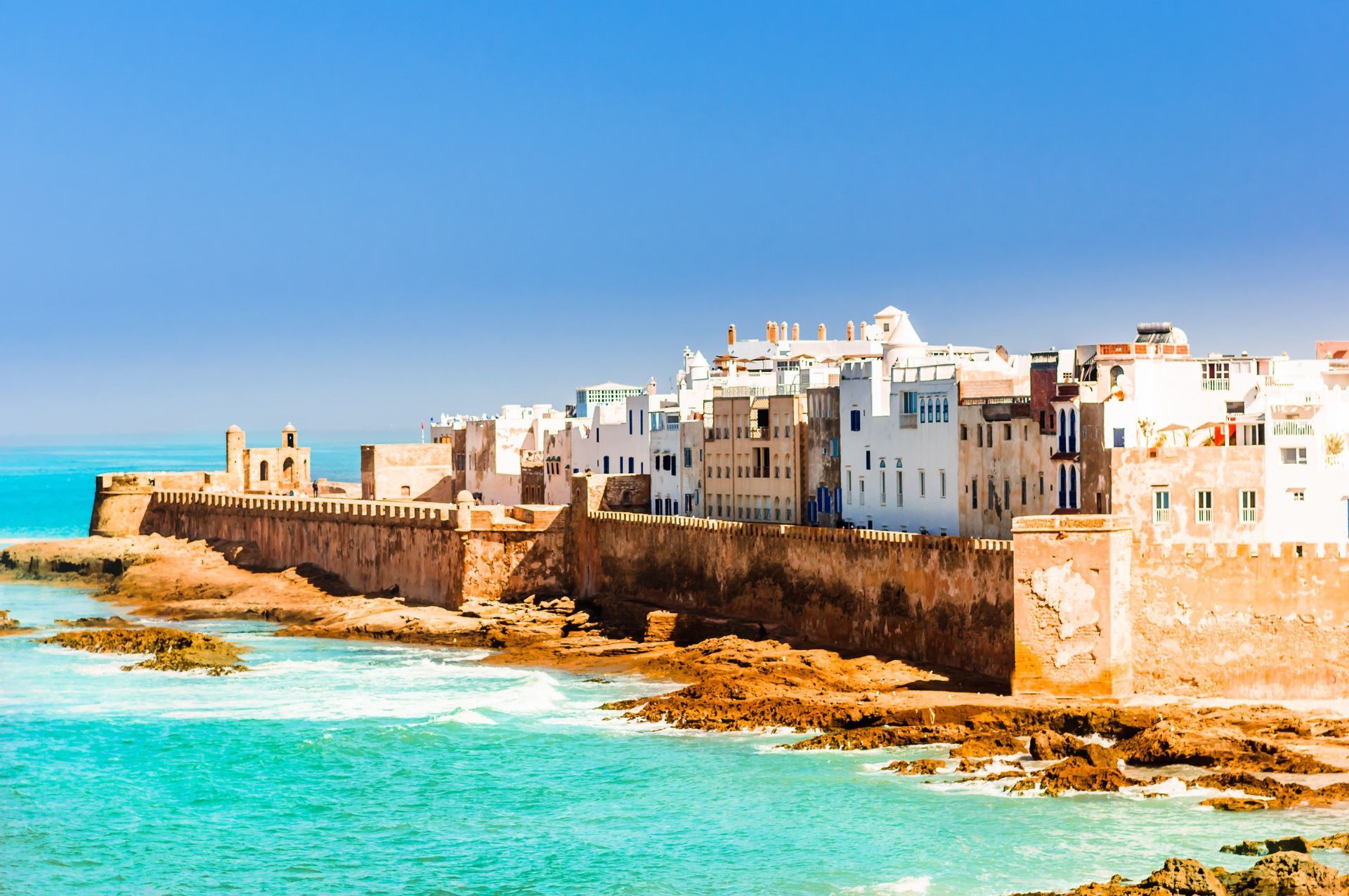 A fortified city with whitewashed buildings on a rocky coast with turquoise water under a clear sky.