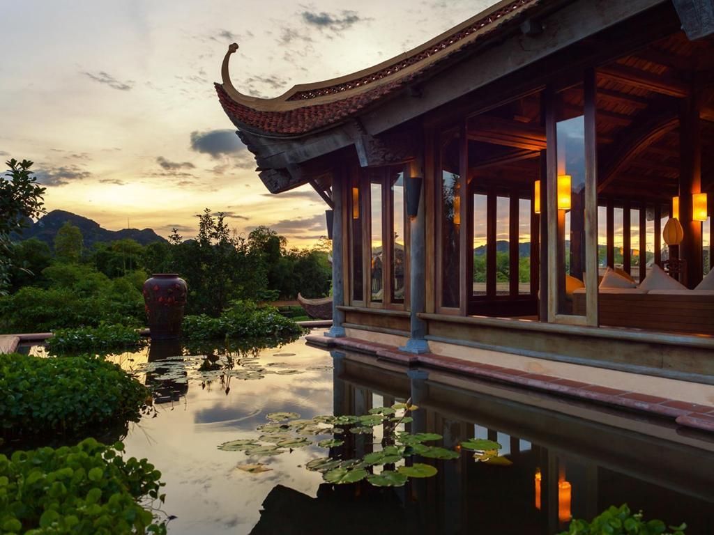 A wooden building with a traditional curved roof overlooks a tranquil pond with water lilies, with lush mountains in the background at sunset.