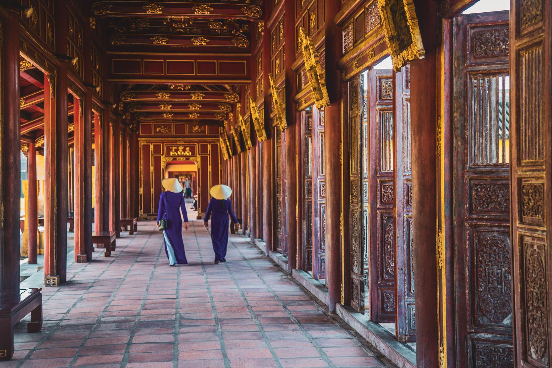 Due donne con cappelli conici e abiti tradizionali viola camminano lungo un lungo e ornato corridoio con pilastri di legno rossi e dorati.
