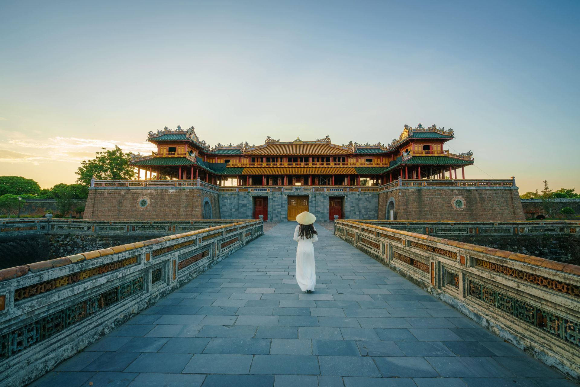 A woman in a white dress and conical hat walks across a stone bridge towards a large, traditional citadel.