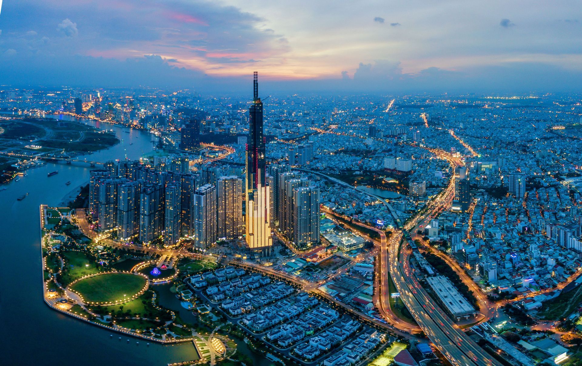 An aerial view of a sprawling metropolis at dusk, with an illuminated central skyscraper, a winding river, and glowing city lights.