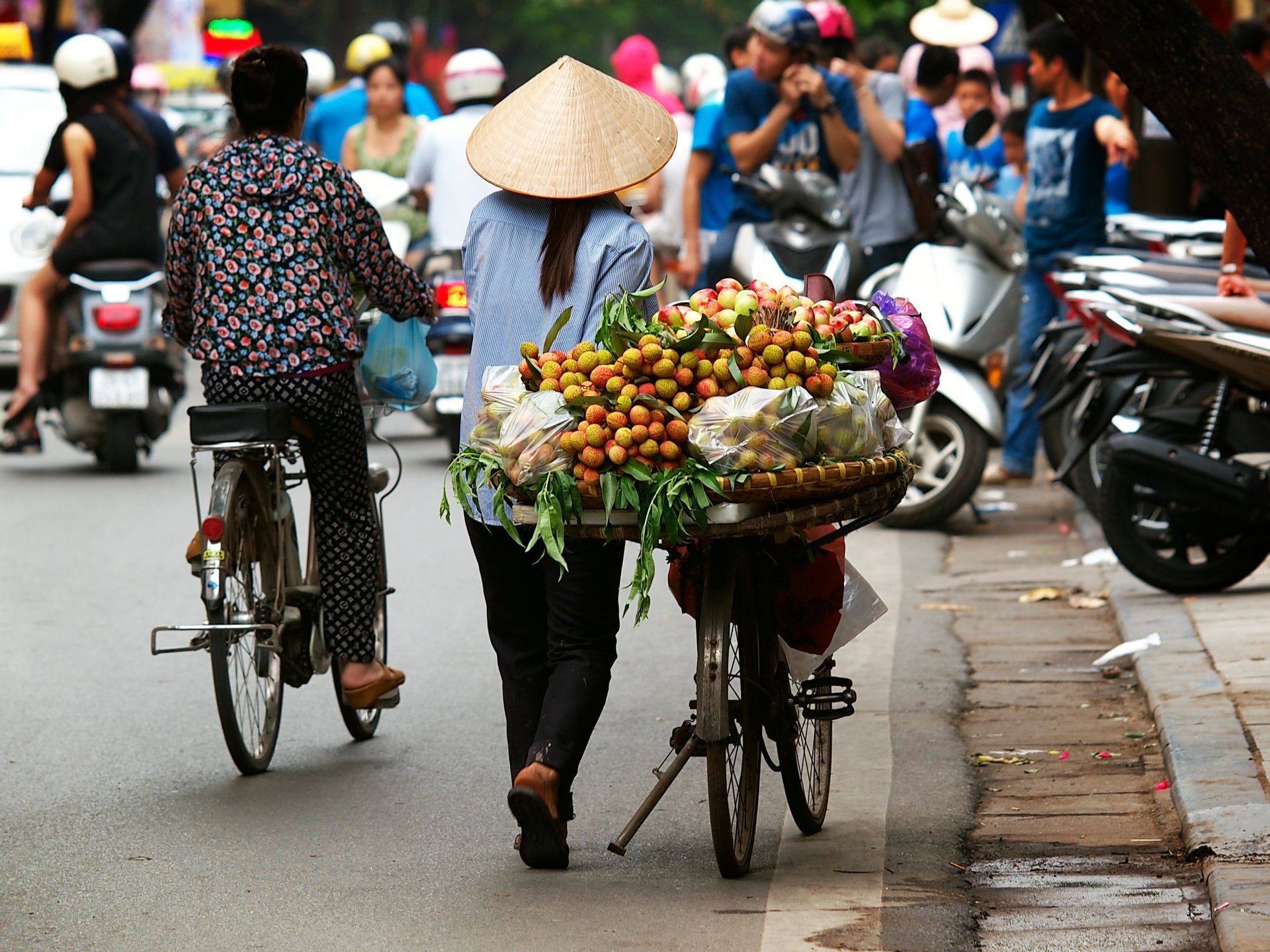 Una persona con un cappello conico spinge una bicicletta carica di cesti di litchi freschi lungo una strada trafficata della città.