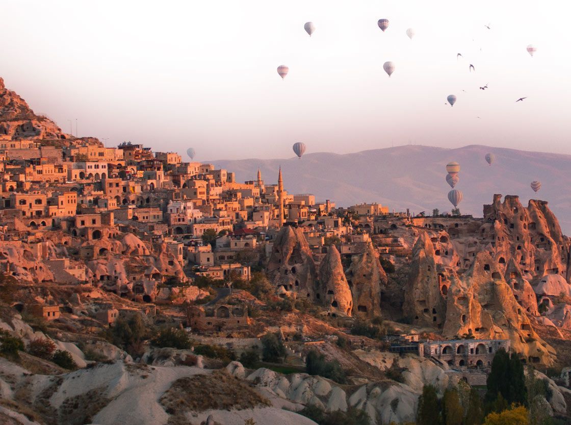 Mongolfiere fluttuano su un villaggio scavato nelle scogliere rocciose e abitazioni rupestri durante un'alba velata.