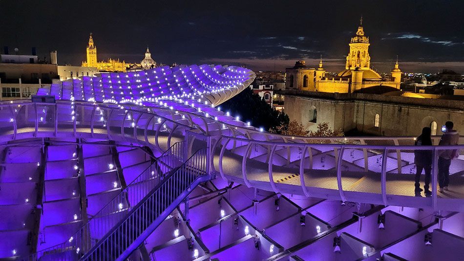 Una struttura architettonica moderna con una passerella sinuosa, illuminata di viola di notte, che si affaccia su una città con edifici storici.