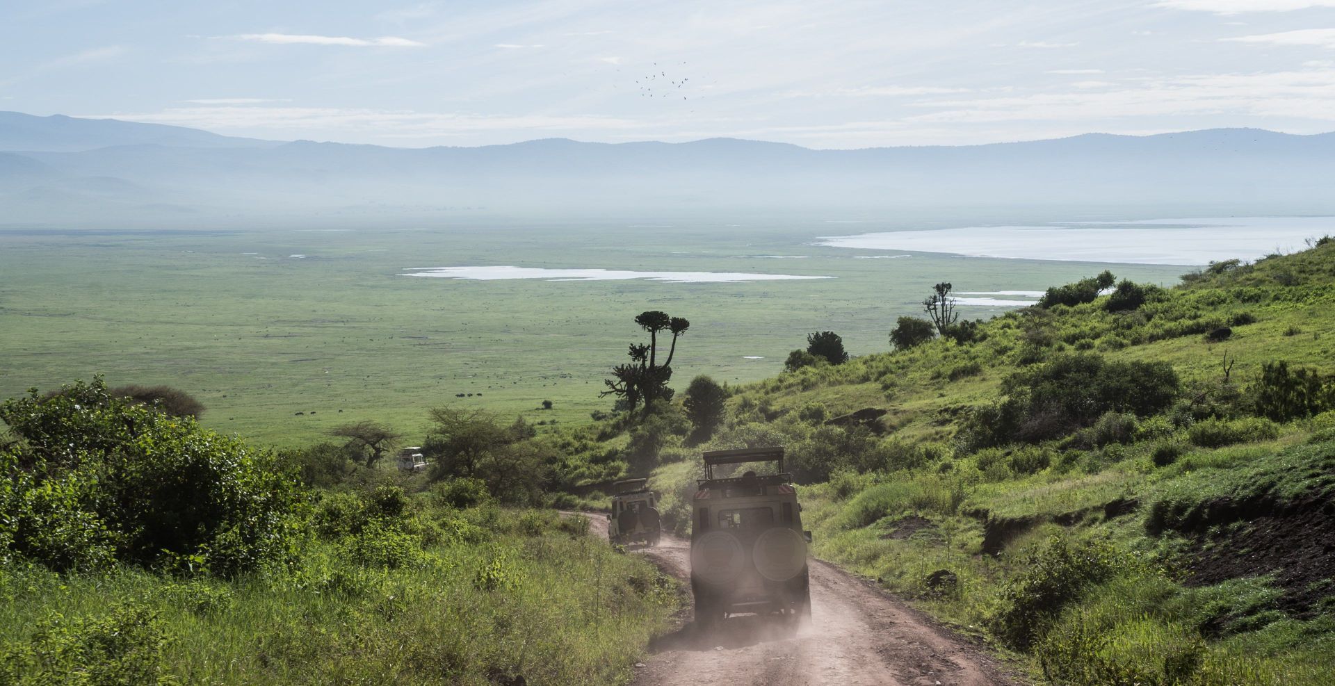 Zwei Safari-Jeeps fahren eine gewundene Schotterstraße einen grasbewachsenen Hügel hinunter und überblicken ein weites grünes Tal mit einem See und fernen Bergen.