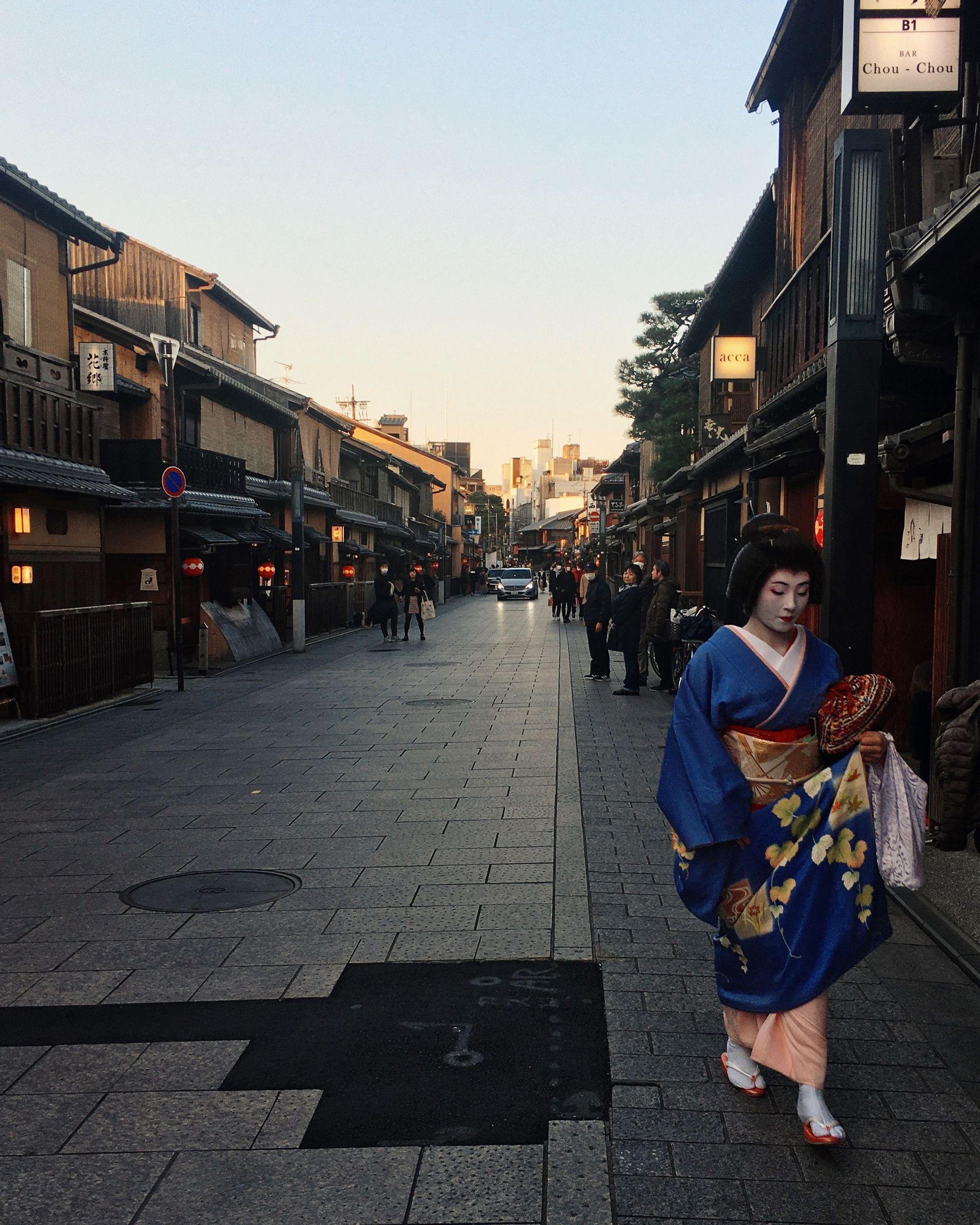 Una donna in un tradizionale kimono blu e trucco bianco percorre una strada lastricata fiancheggiata da edifici tradizionali in legno al crepuscolo.