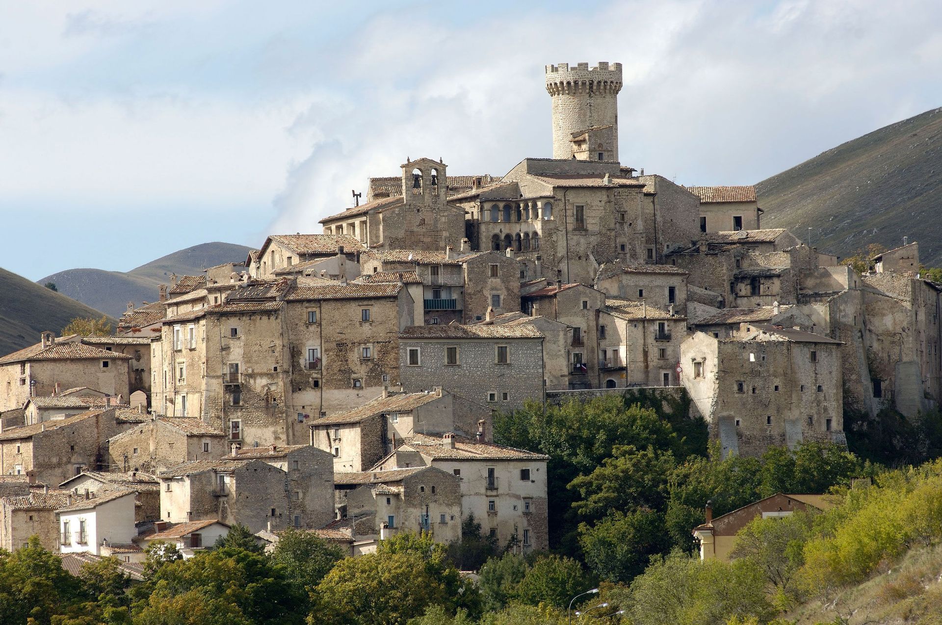 Un antico borgo in pietra con una torre rotonda costruito su una verde collina sotto un cielo parzialmente nuvoloso.