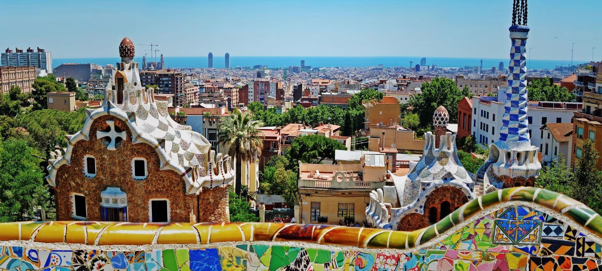 Edifici fantasiosi, rivestiti a mosaico, che si affacciano su un vasto panorama urbano che incontra il mare sotto un cielo azzurro e limpido.