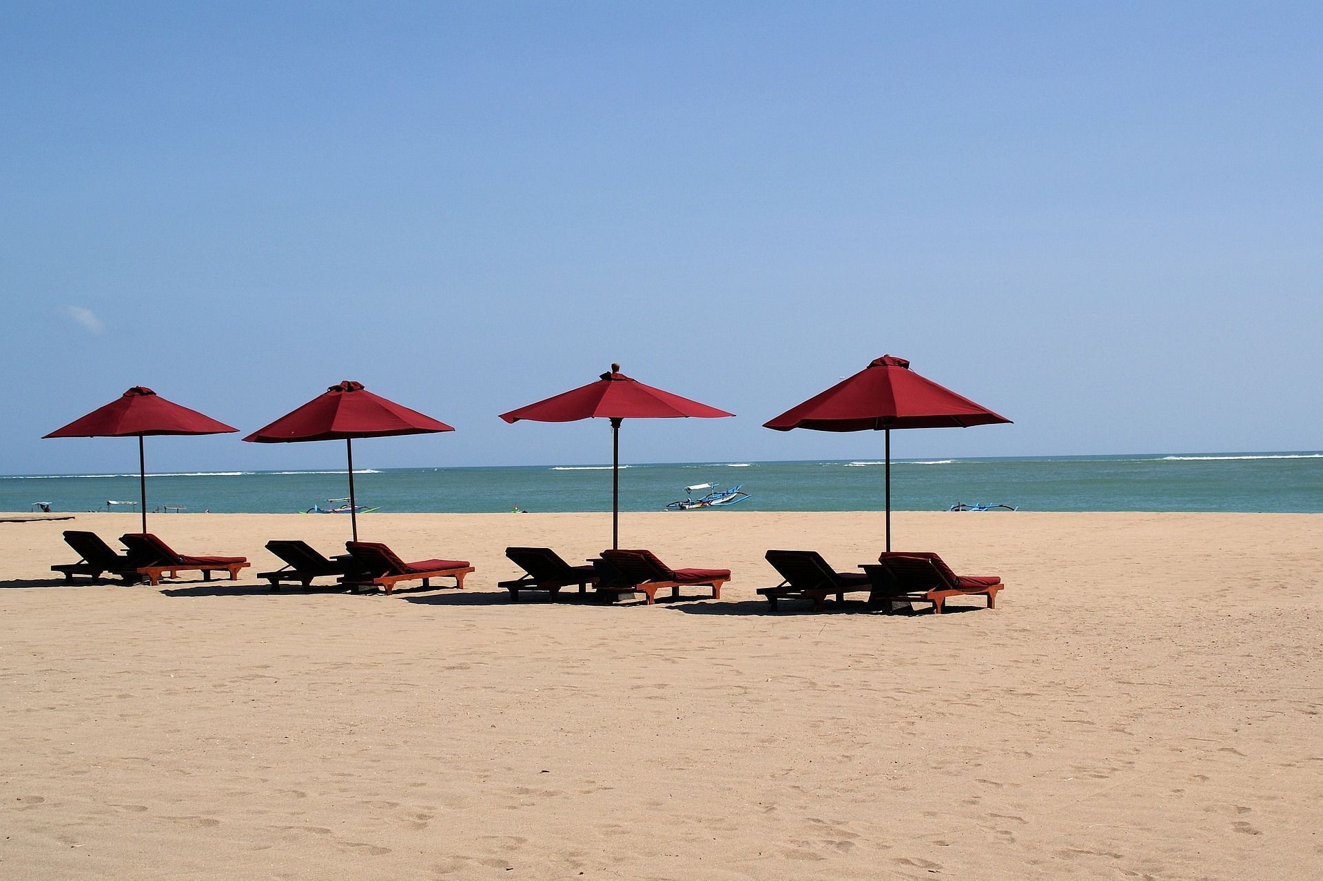 Une rangée de parasols rouges et de transats vides sur une plage de sable fin face à l'océan, sous un ciel dégagé.
