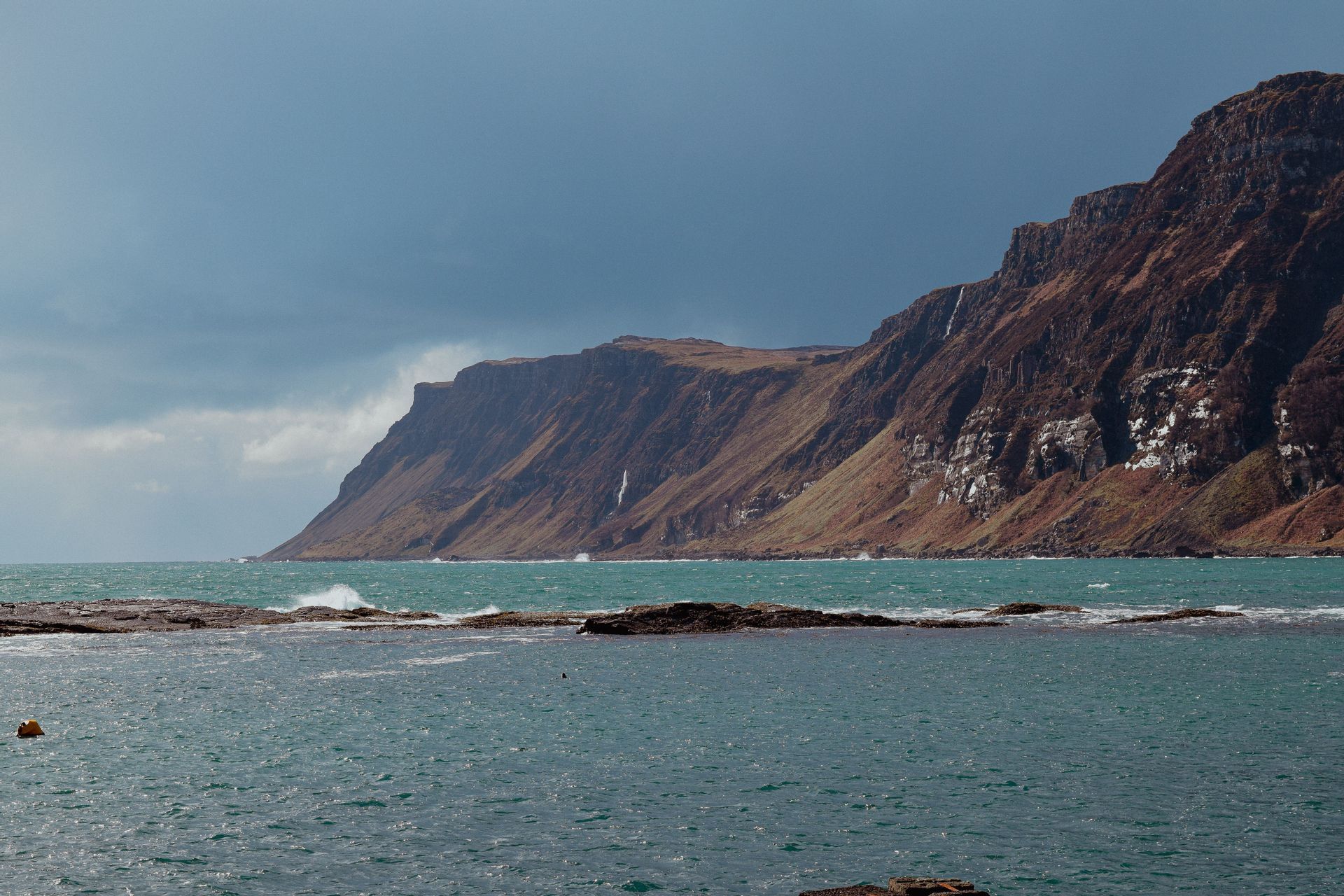 A rugged, brown cliffside with several waterfalls overlooks a choppy turquoise sea under an overcast sky.