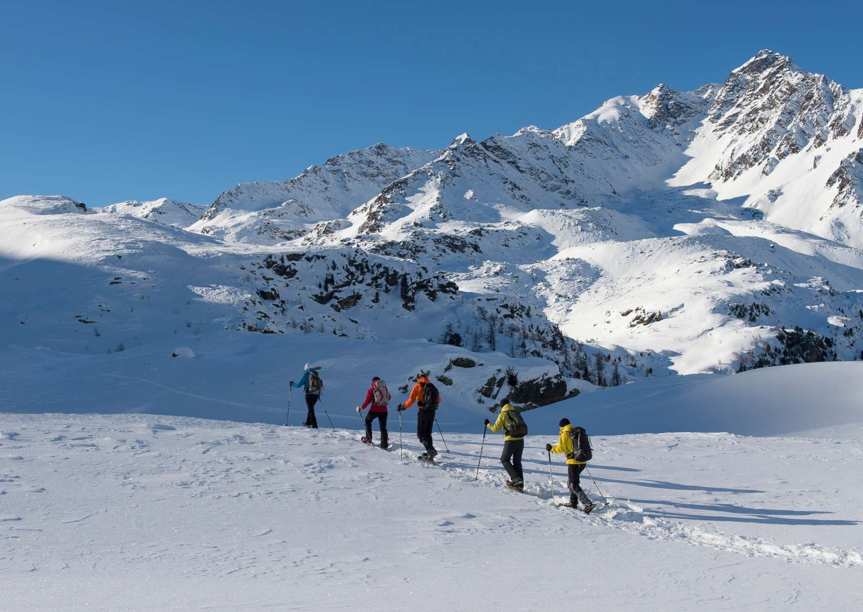Un viaggio di gruppo WeRoad con ciaspolata in fila indiana, circondati da montagne innevate sotto un cielo sereno.