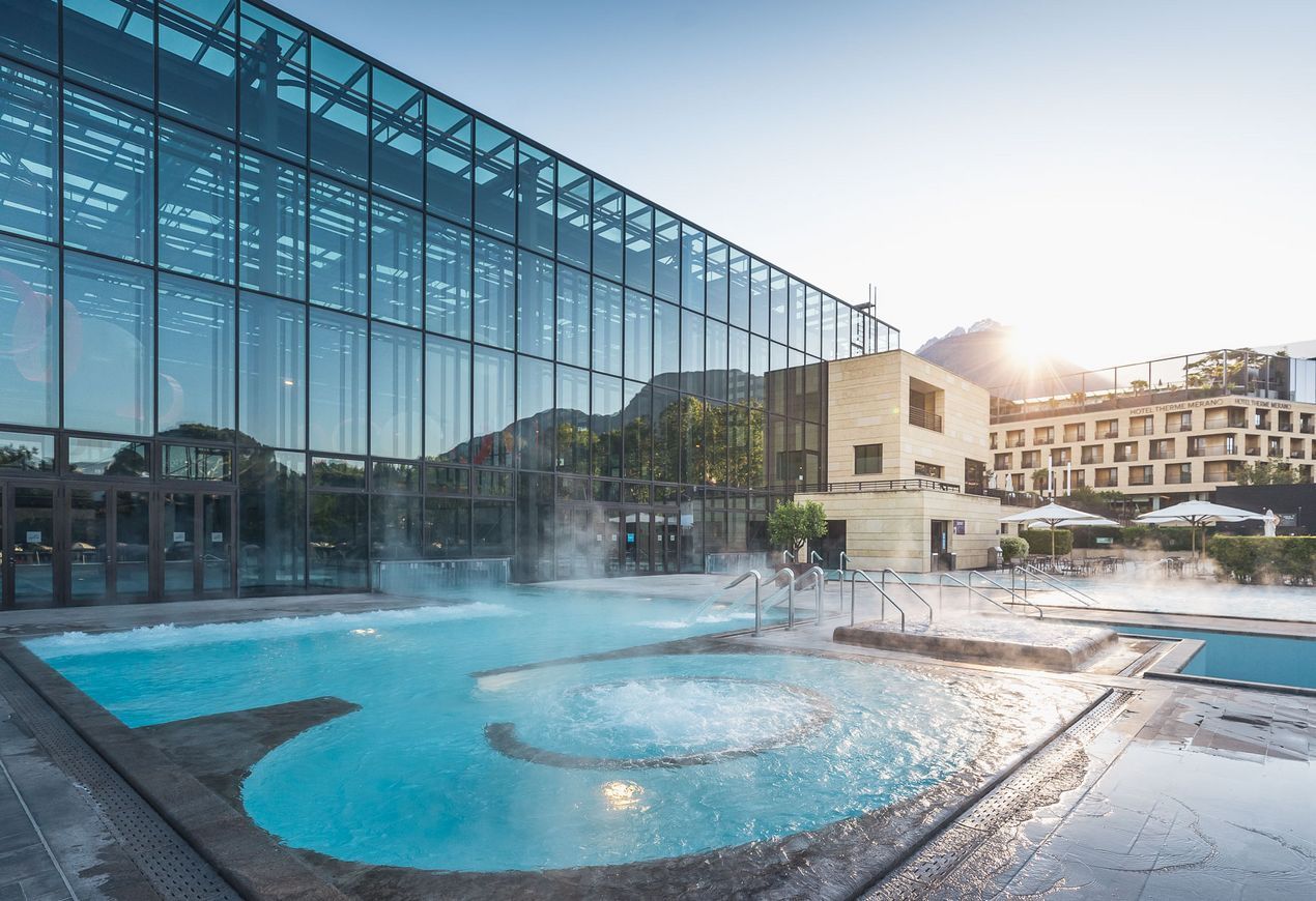 Piscina termale all'aperto con vapore che sale dall'acqua, accanto a un moderno edificio con facciata in vetro all'alba.