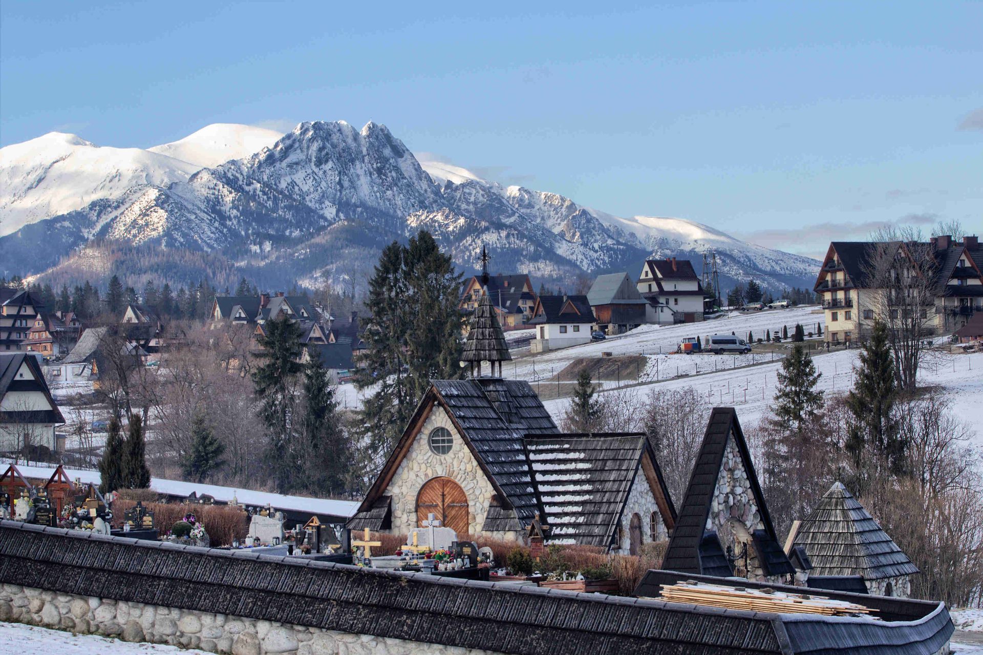 A stone chapel and village in a snowy valley at the foot of a large, snow-capped mountain range.