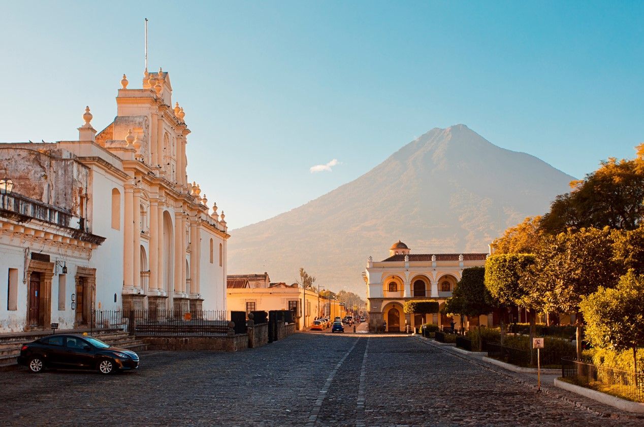 Una vista su una strada acciottolata fiancheggiata da edifici coloniali, con un grande vulcano in lontananza sotto un cielo azzurro chiaro.