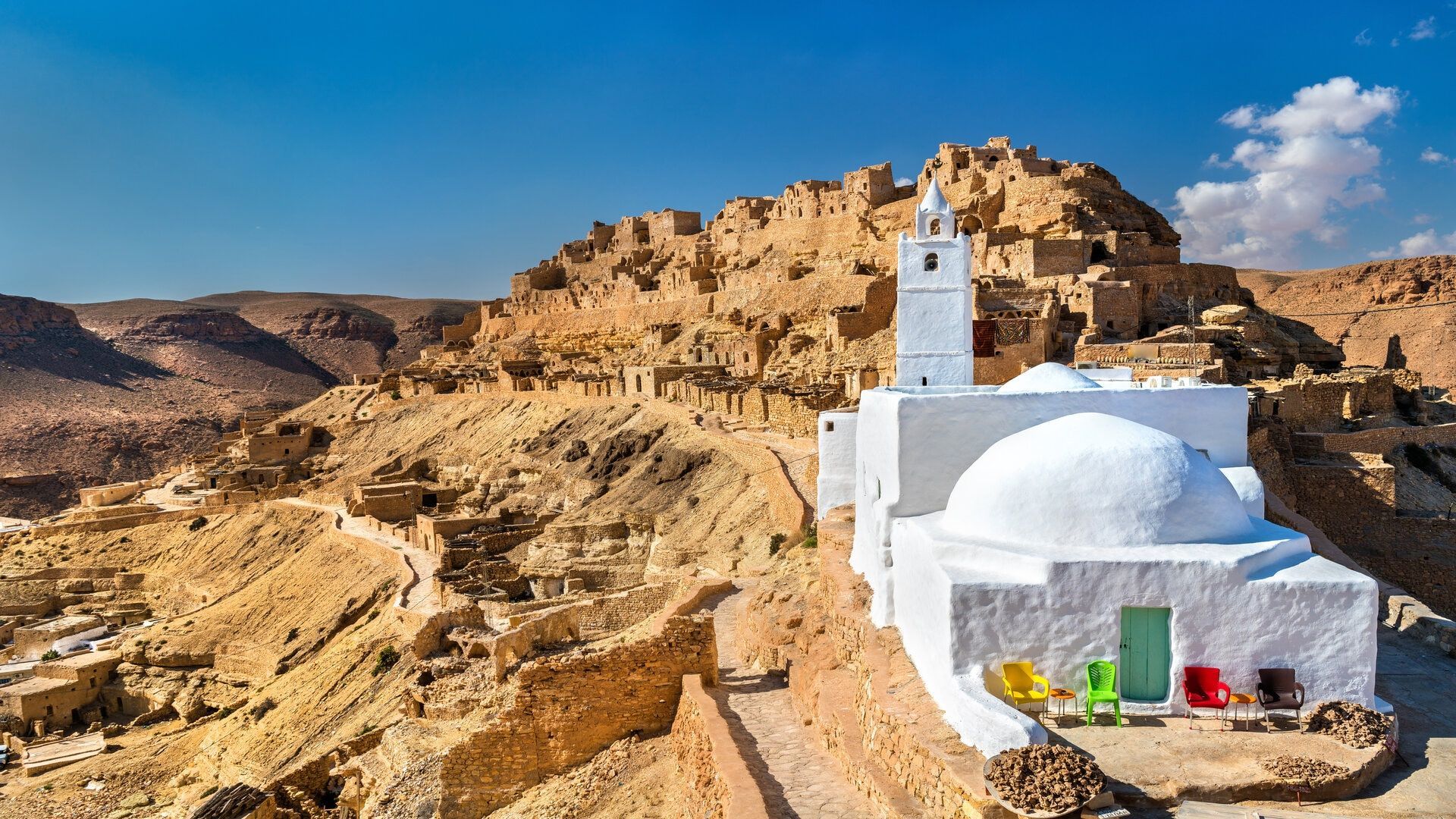 Un edificio imbiancato a calce con un minareto si erge in un villaggio desertico, incastonato in una collina rocciosa e assolata, sotto un cielo azzurro e limpido.