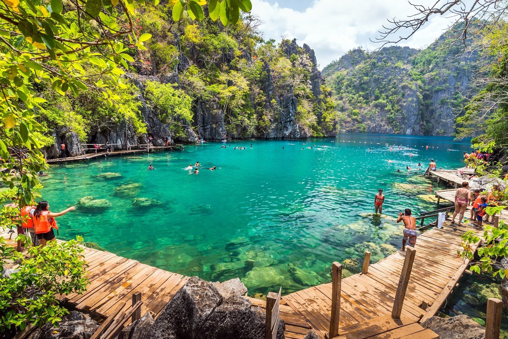 A WeRoad group trip swimming in a clear turquoise lagoon surrounded by rocky cliffs with lush greenery and wooden walkways.