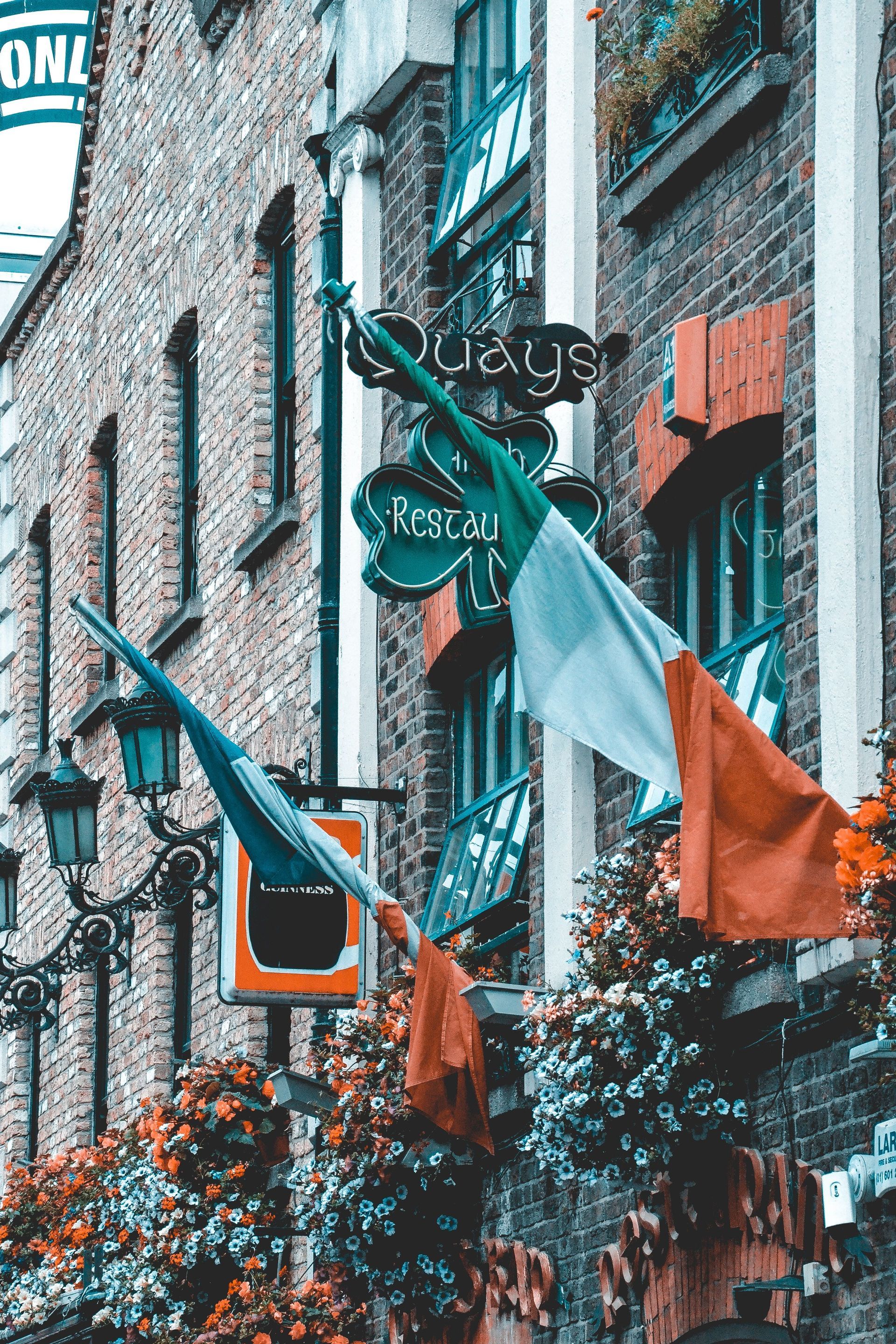 Una bandera irlandesa ondea frente a la fachada de ladrillo de un pub, adornada con coloridas flores y carteles antiguos.