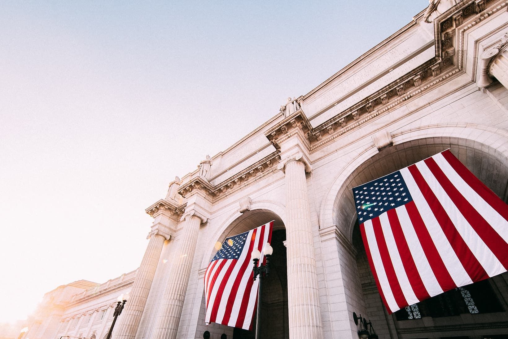 A low-angle view of two large American flags hanging from the arches of a classical stone building.