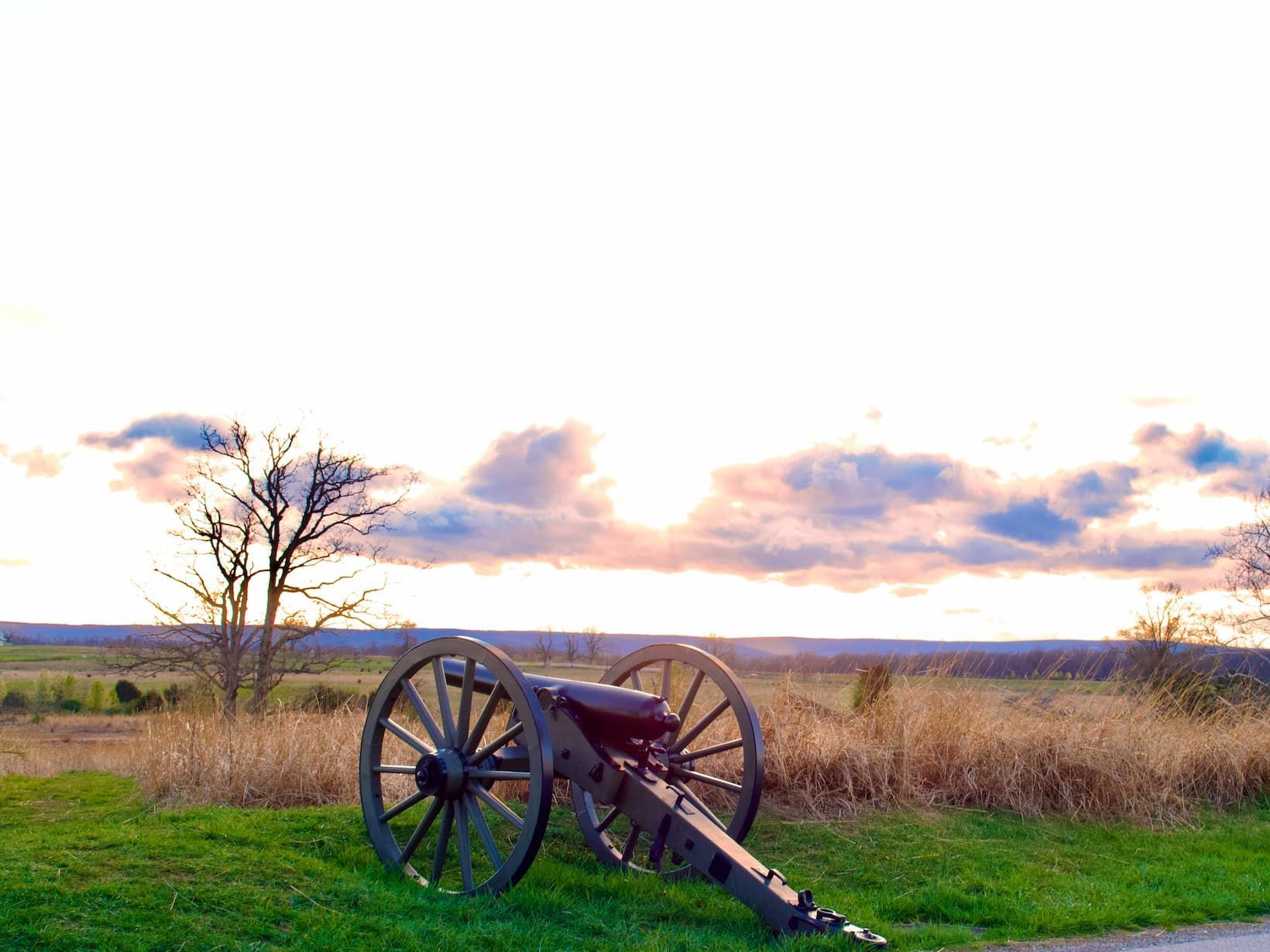 A historic cannon sits on a grassy hill overlooking a field with a bare tree under a cloudy sunset sky.