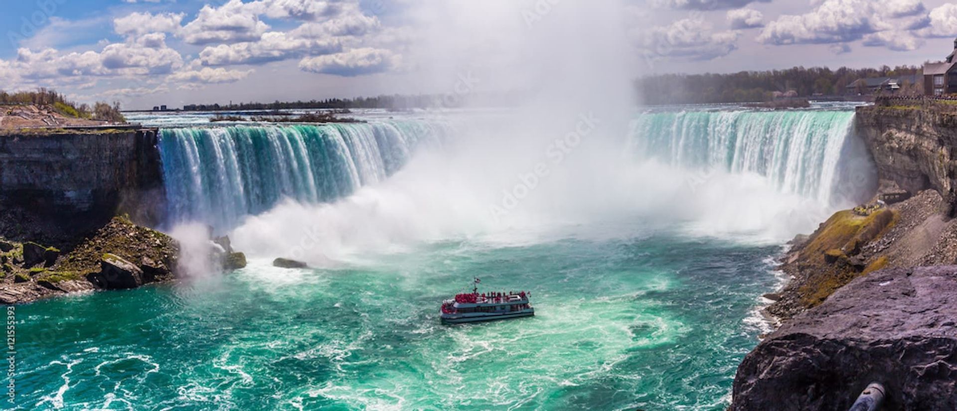 A tour boat sails on turbulent turquoise water near the base of a massive, horseshoe-shaped waterfall under a cloudy sky.