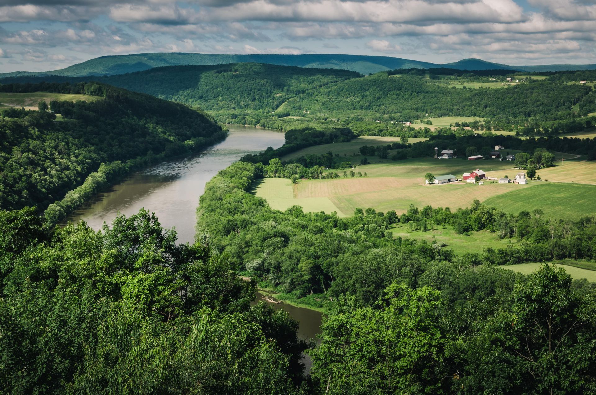 An elevated view of a river winding through a valley of green forests and fields, with rolling hills in the distance under a cloudy sky.