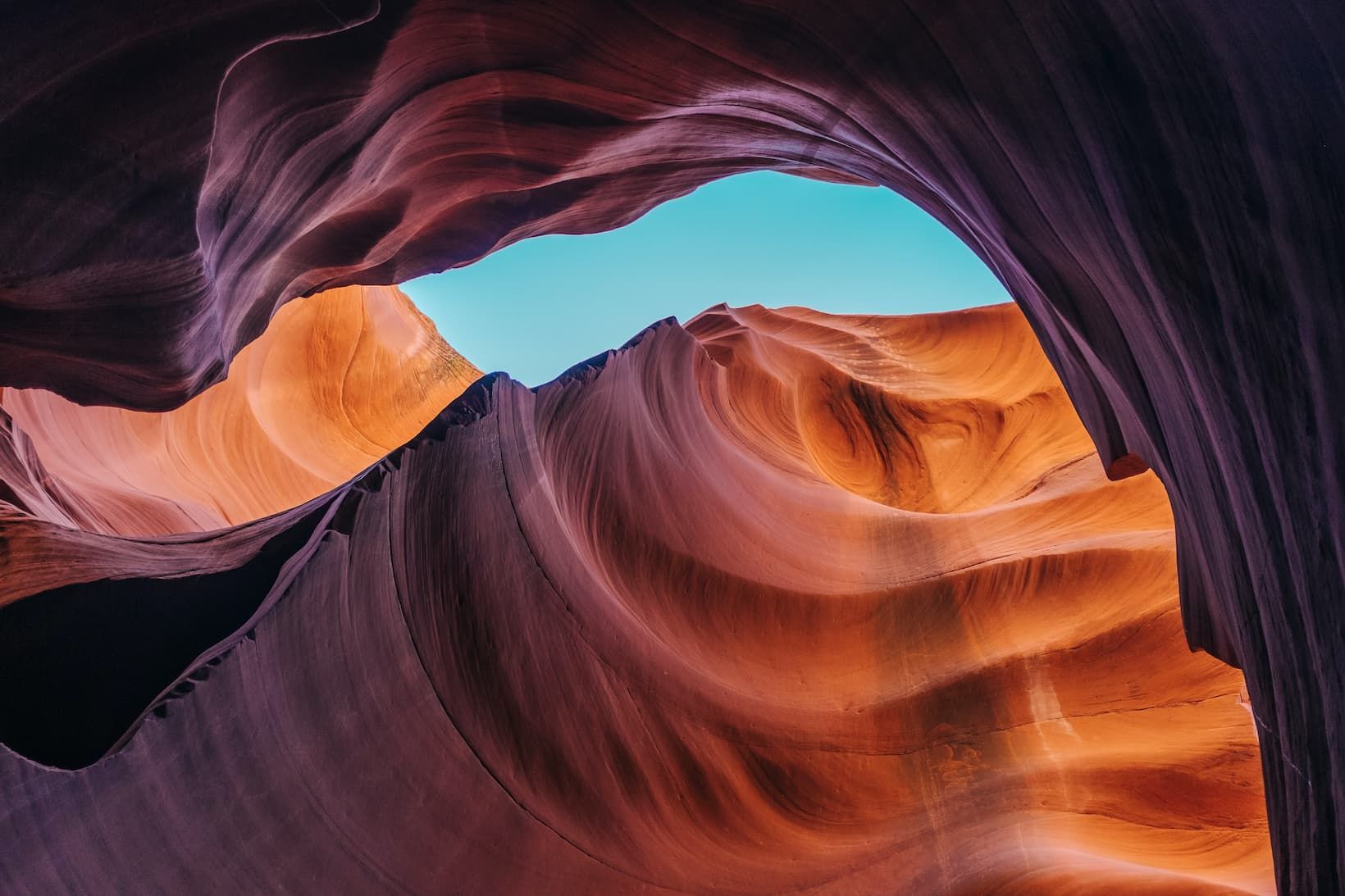 Formazioni di arenaria ondulate e stratificate di un canyon a fessura, nelle tonalità arancioni e viola, che incorniciano uno squarcio di cielo azzurro limpido.