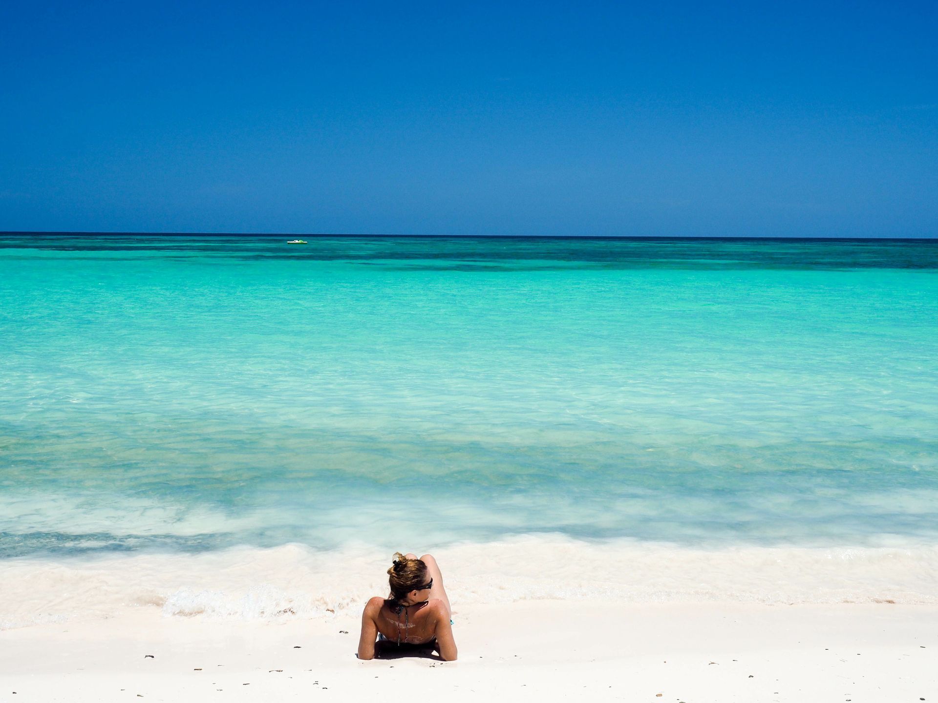 Una donna con i capelli raccolti a chignon giace su una spiaggia di sabbia bianca, guardando il mare calmo e turchese.