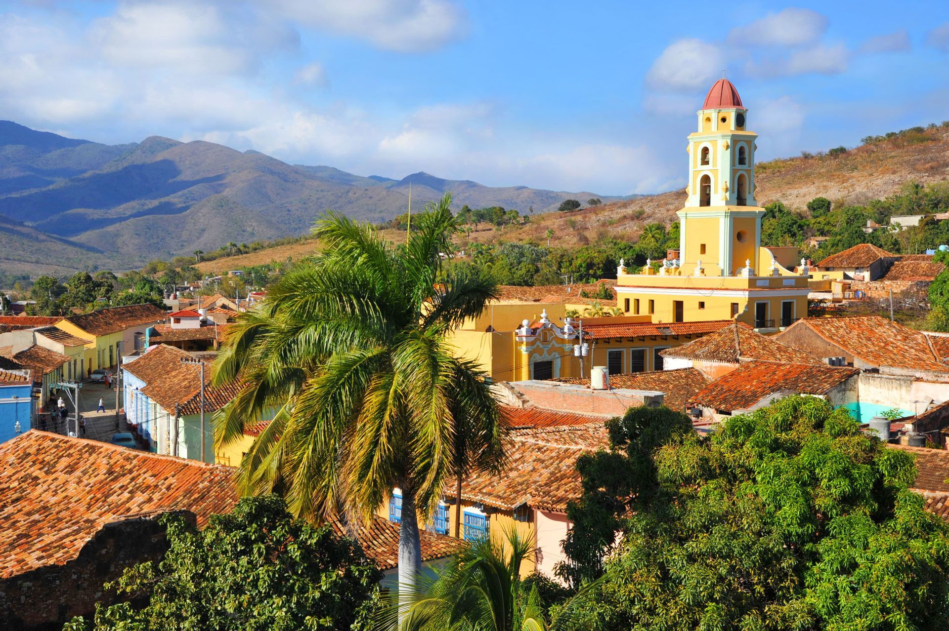 Un campanile giallo si erge su una città con tetti in terracotta, con palme e montagne verdi sullo sfondo.