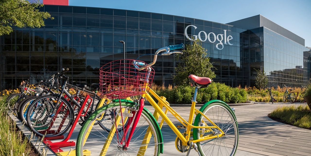 Biciclette colorate parcheggiate in una rastrelliera davanti alla moderna sede in vetro di Google in una giornata di sole.