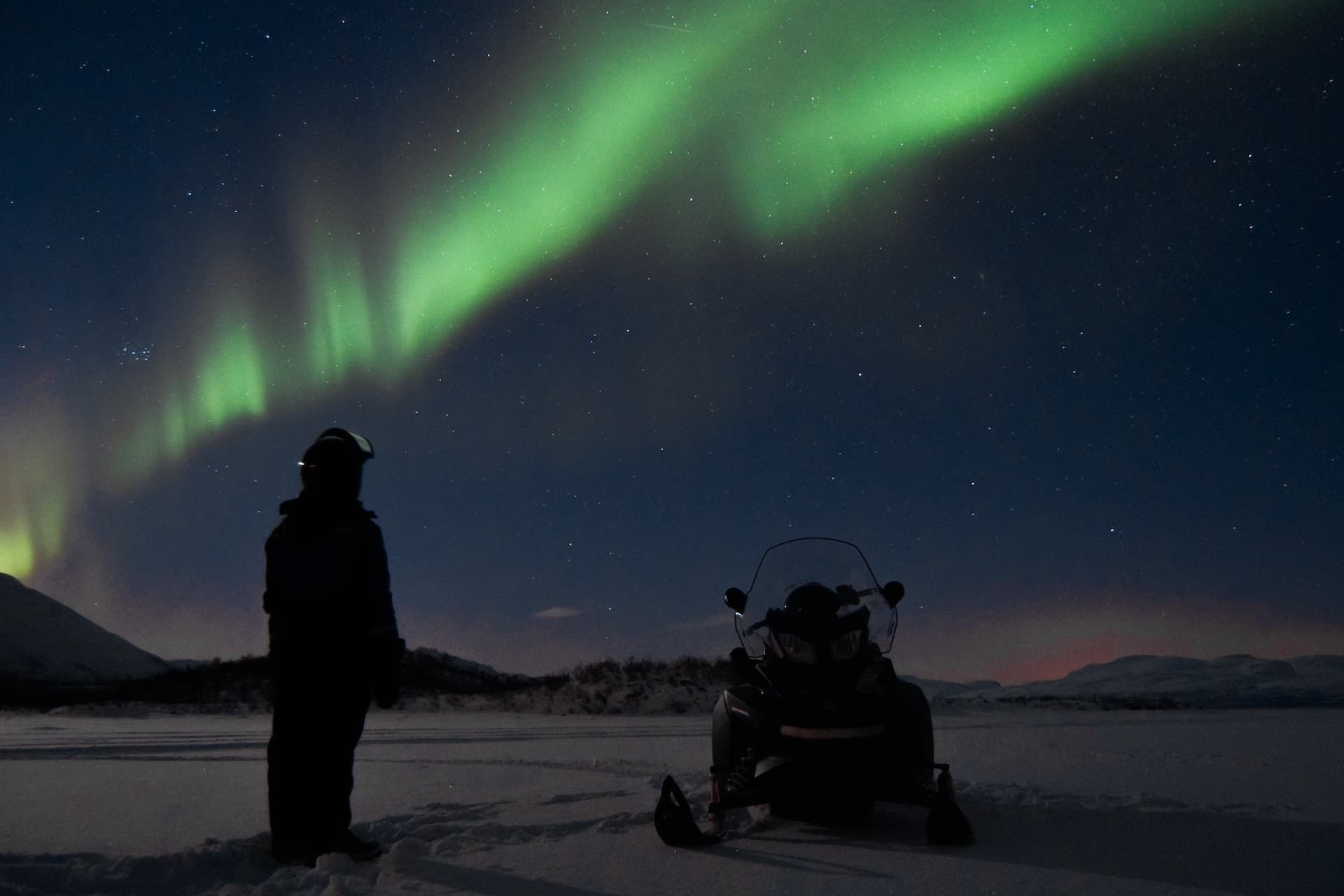 Una persona junto a una moto de nieve en un paisaje nevado nocturno, contemplando la aurora boreal verde que llena el cielo estrellado.