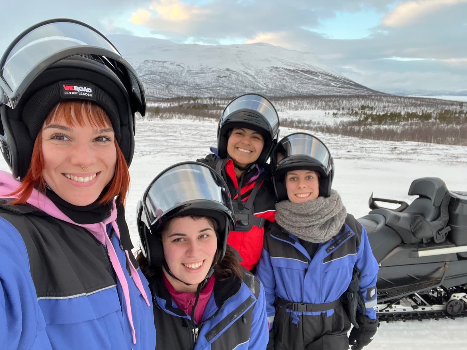 A WeRoad group trip of four women wearing helmets smiles for a selfie next to a snowmobile in a snowy, mountainous landscape.