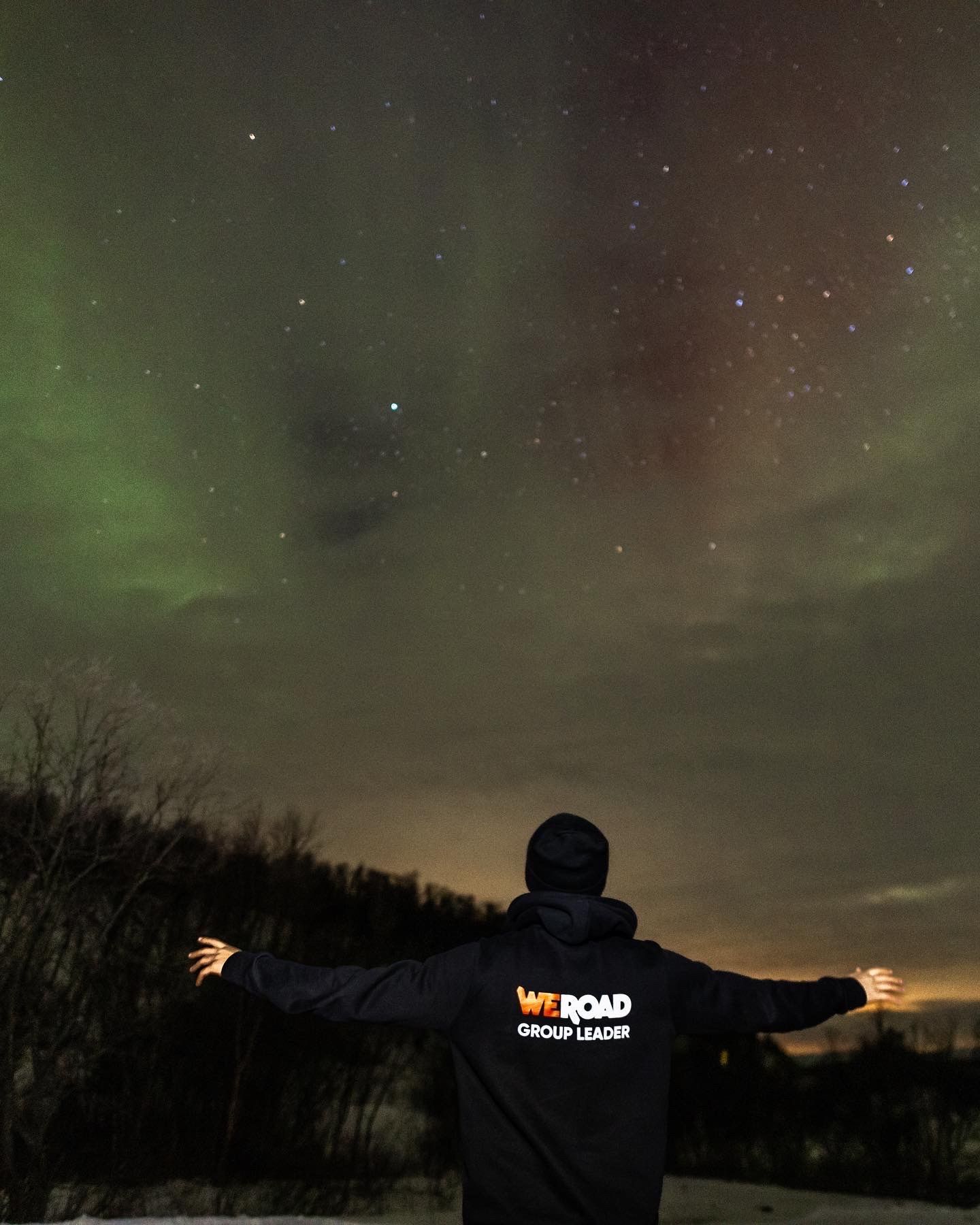 A WeRoad Group Leader stands with arms outstretched, looking up at the aurora borealis and starry night sky.