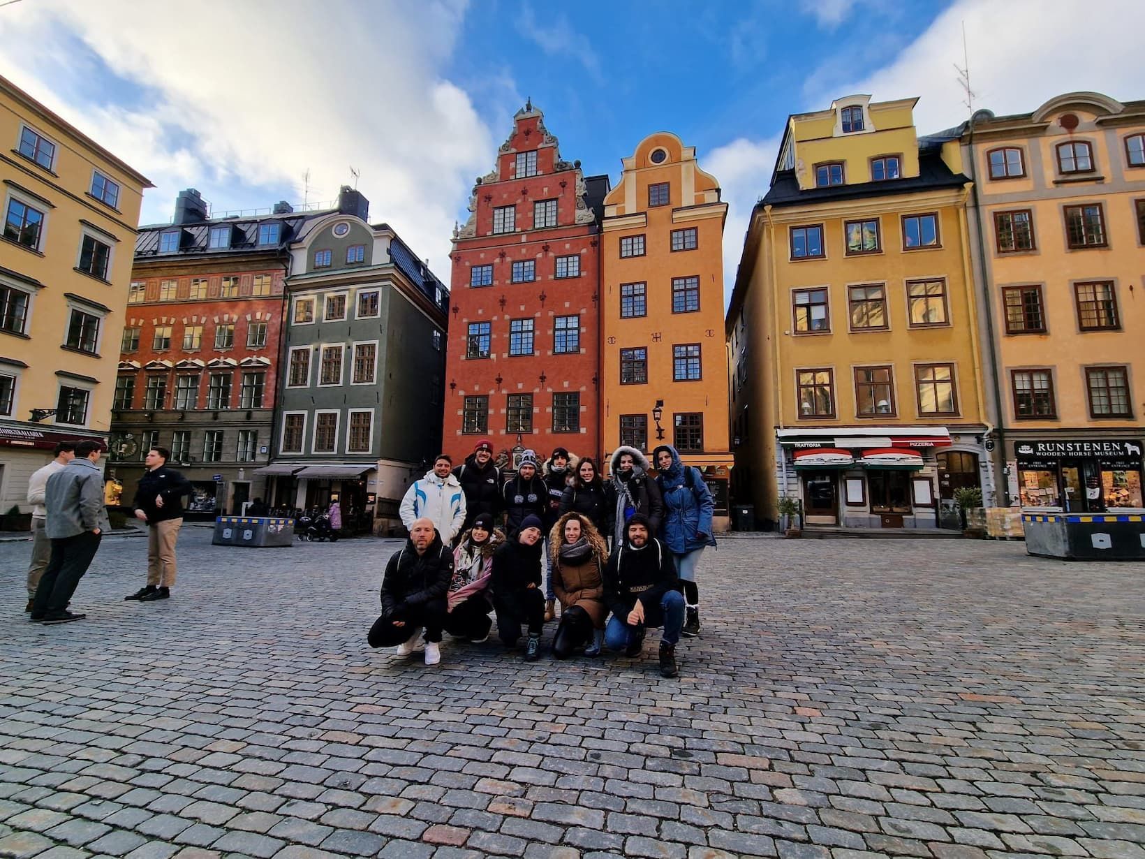 A WeRoad group trip poses for a photo in a cobblestone square in front of colorful historic buildings.