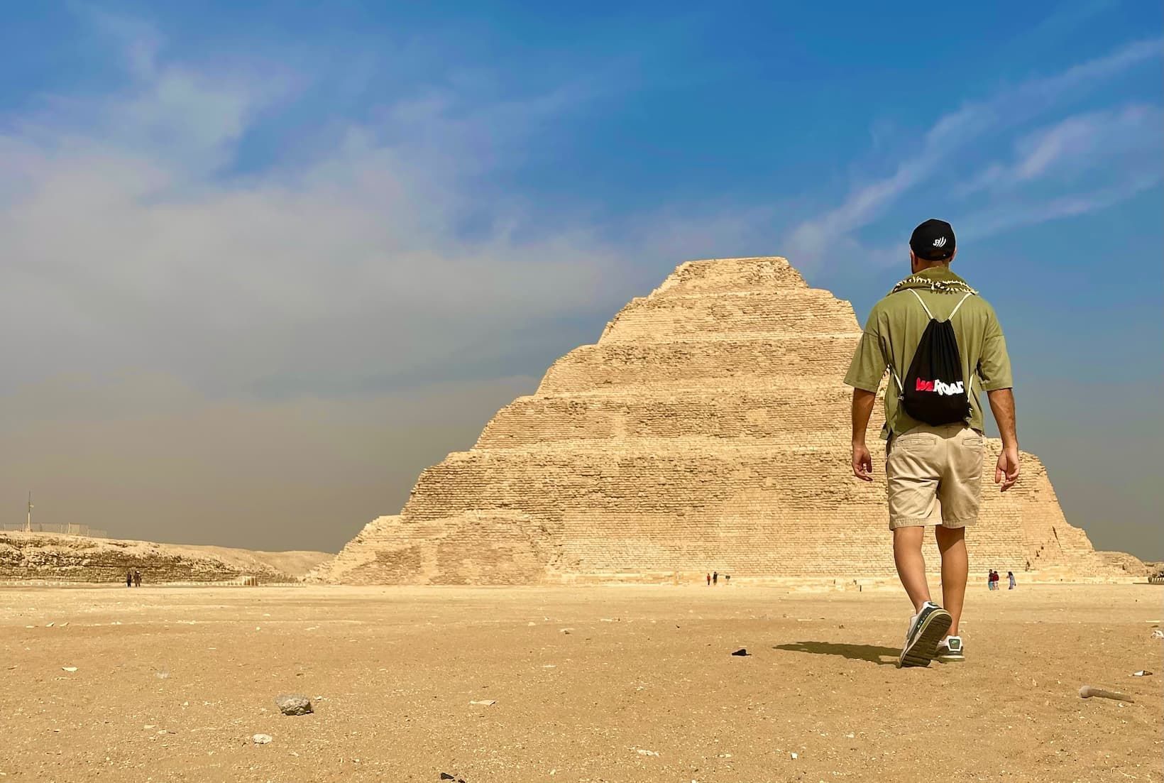 A traveler with a WeRoad backpack walks across a sandy desert landscape towards a large step pyramid.