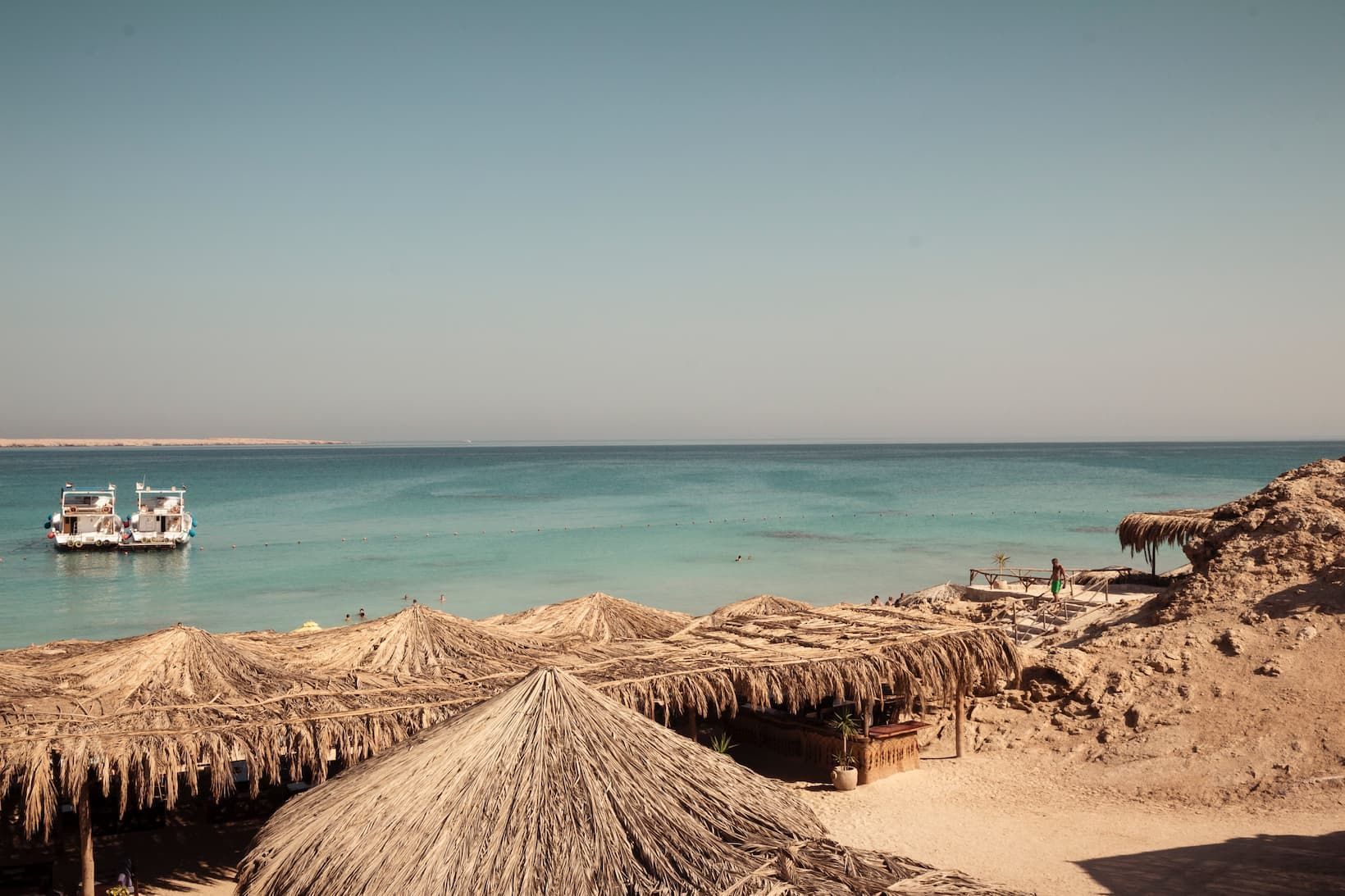 Parasols au toit de chaume sur une plage de sable avec deux bateaux ancrés dans une eau calme et turquoise sous un ciel clair.