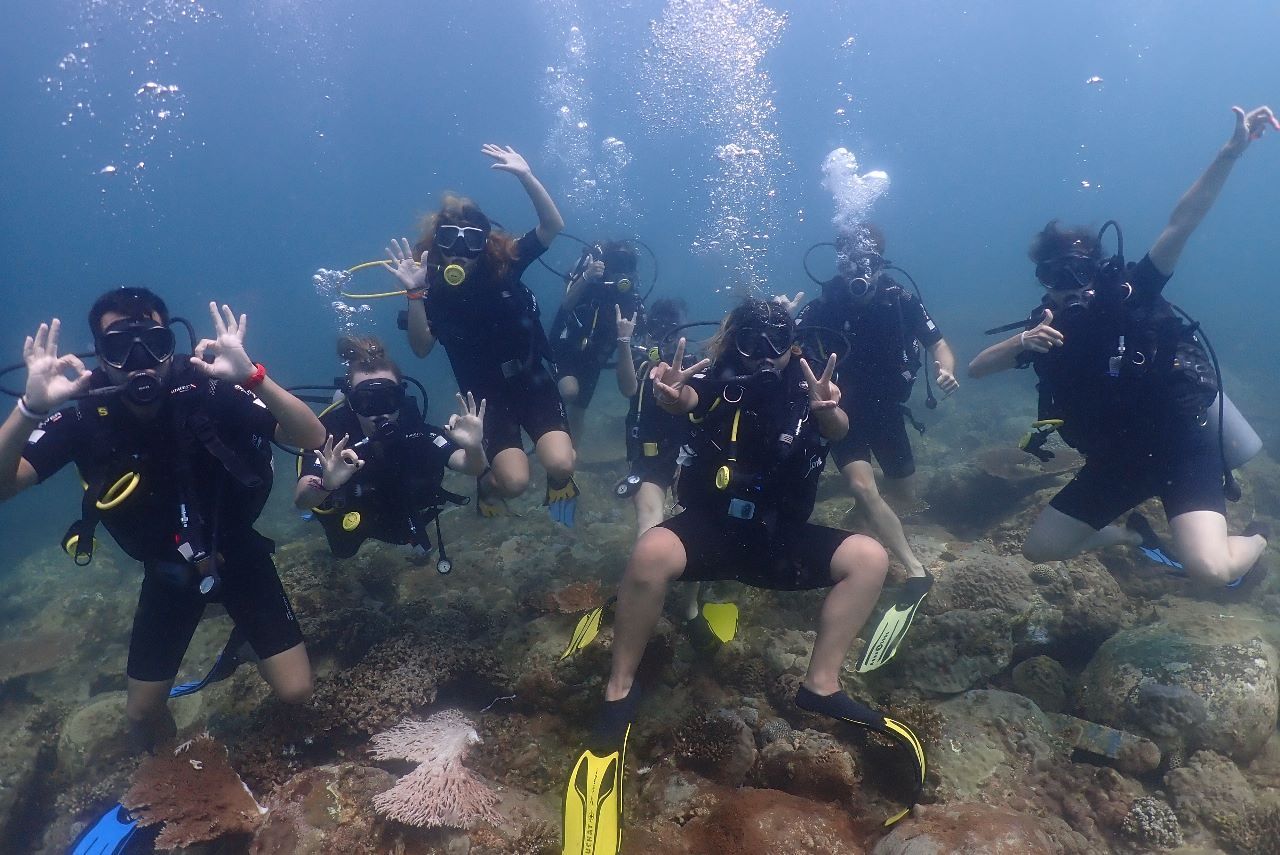 Un gruppo WeRoad in attrezzatura subacquea posa per una foto sottomarina, facendo segni con le mani sopra una scogliera rocciosa.