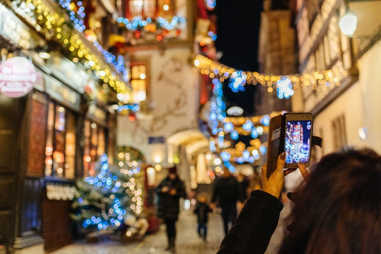 A person holds up a smartphone to photograph a narrow street decorated with colorful Christmas lights at night.
