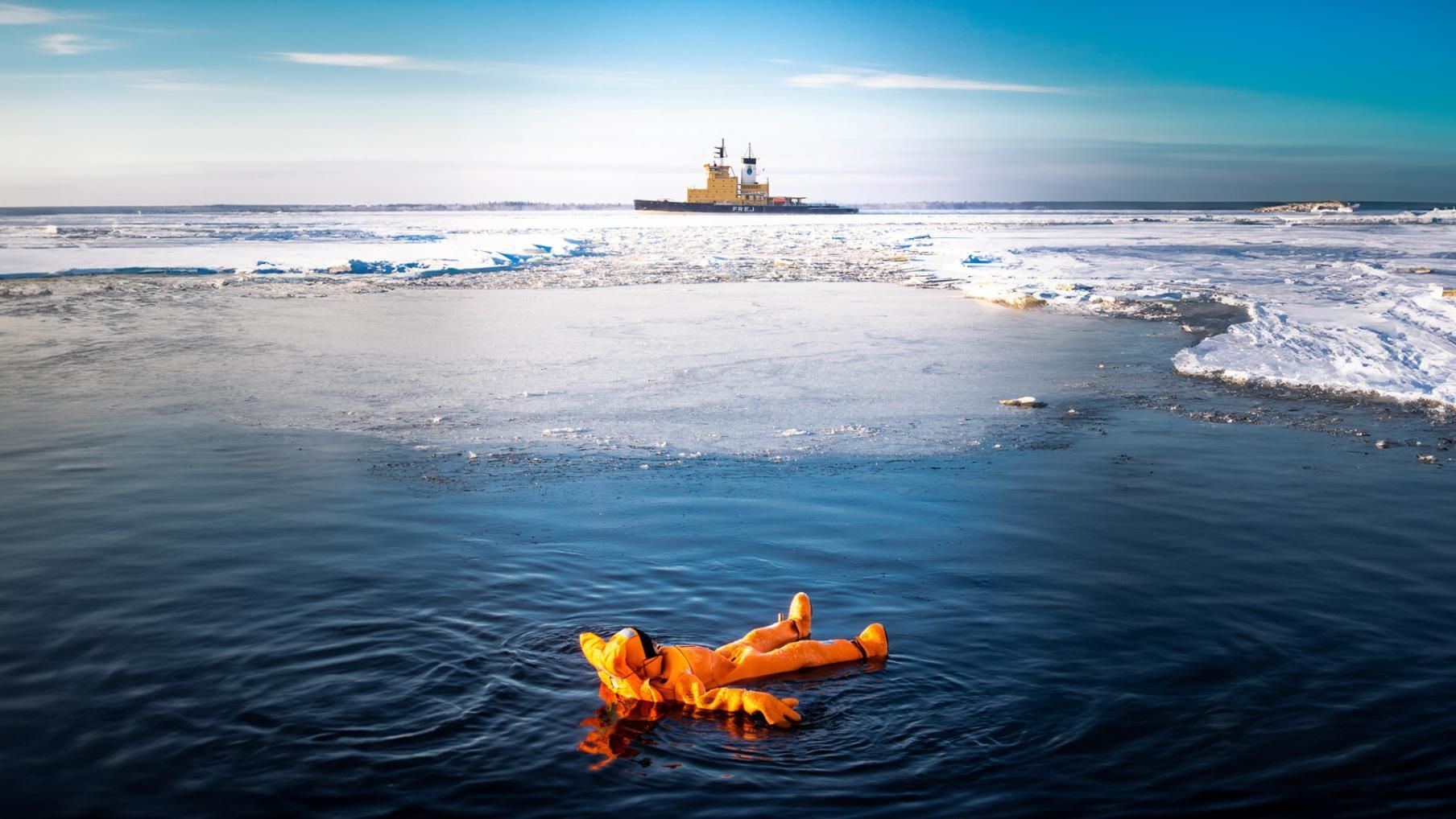 Une personne en combinaison de survie orange flotte sur le dos dans l'eau glacée, avec un brise-glace au loin.