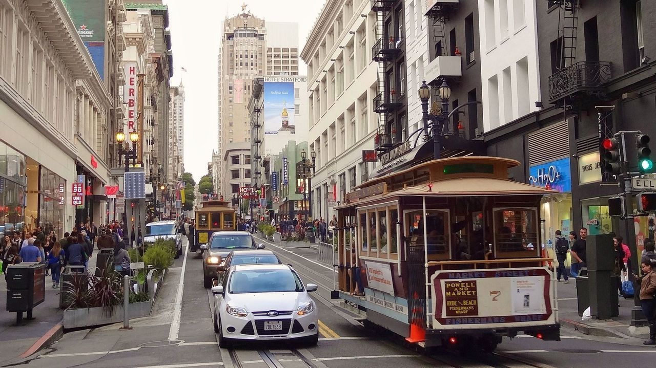 Un cable car di San Francisco percorre una trafficata strada cittadina, condividendo la carreggiata con le auto mentre i pedoni camminano sui marciapiedi.