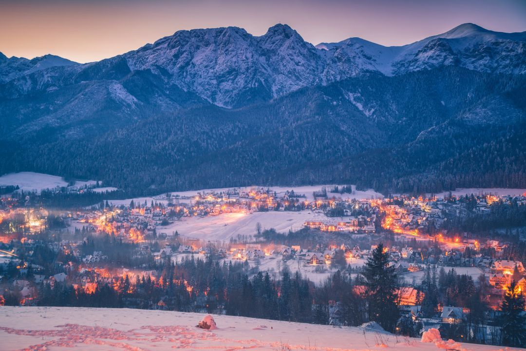 A snow-covered village with glowing lights nestled in a valley at the foot of a large mountain range at dusk.