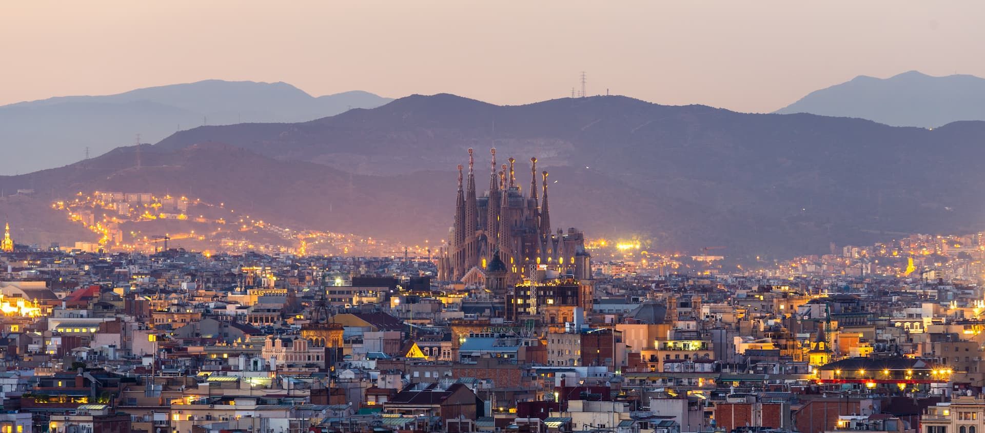 An illuminated cityscape featuring a large ornate cathedral, with a mountain range in the background at dusk.