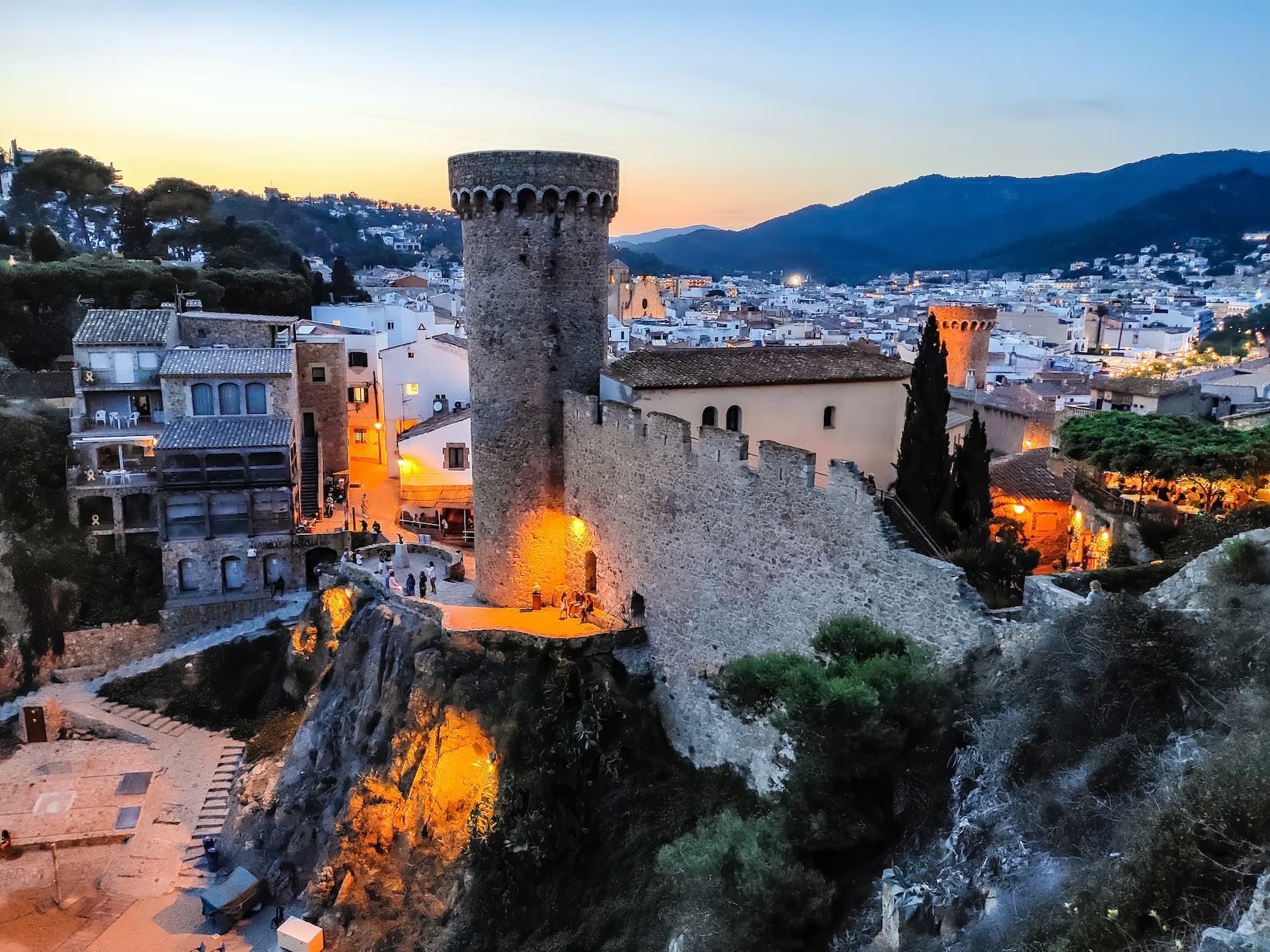 Un château de pierre avec une tour est illuminé sur une falaise dominant une ville et des montagnes au crépuscule.